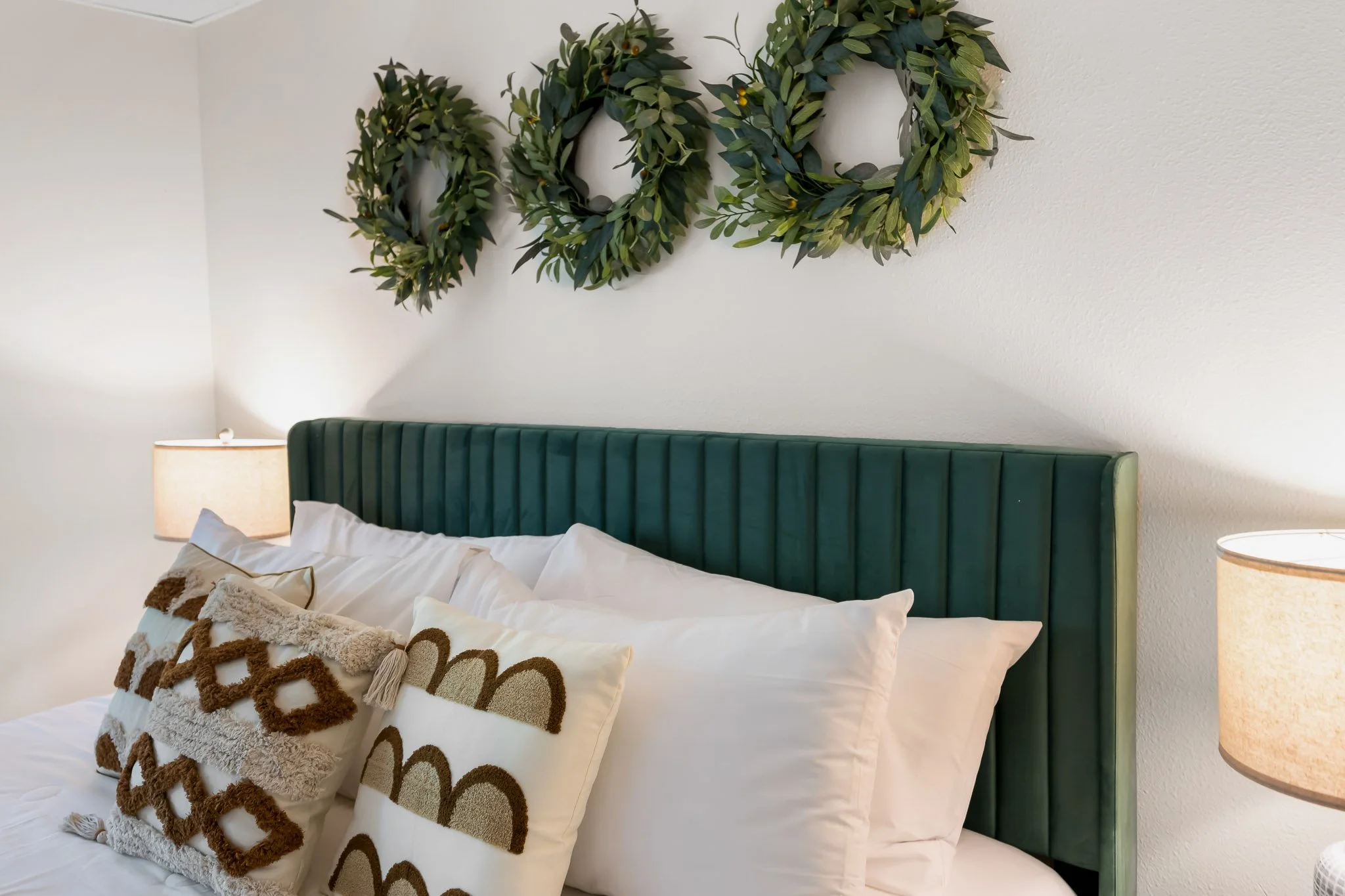 A bedroom with a green upholstered headboard, white bedding, decorative pillows, a nightstand with a lamp, and a green leafy wreath hanging on a white wall.