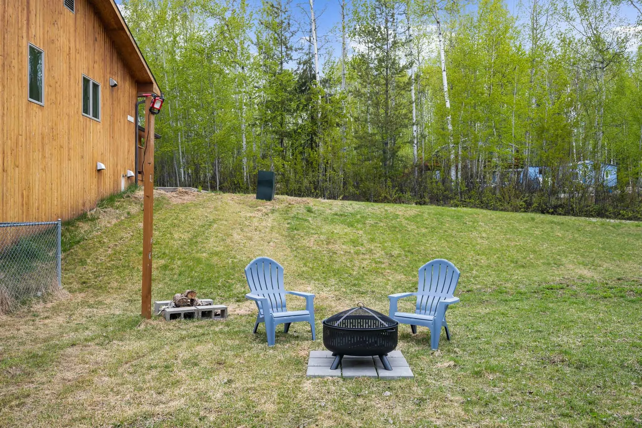 A backyard with two blue plastic chairs, a fire pit, and a firewood storage area. There's a wooden house on the left and a forest of green trees in the background.