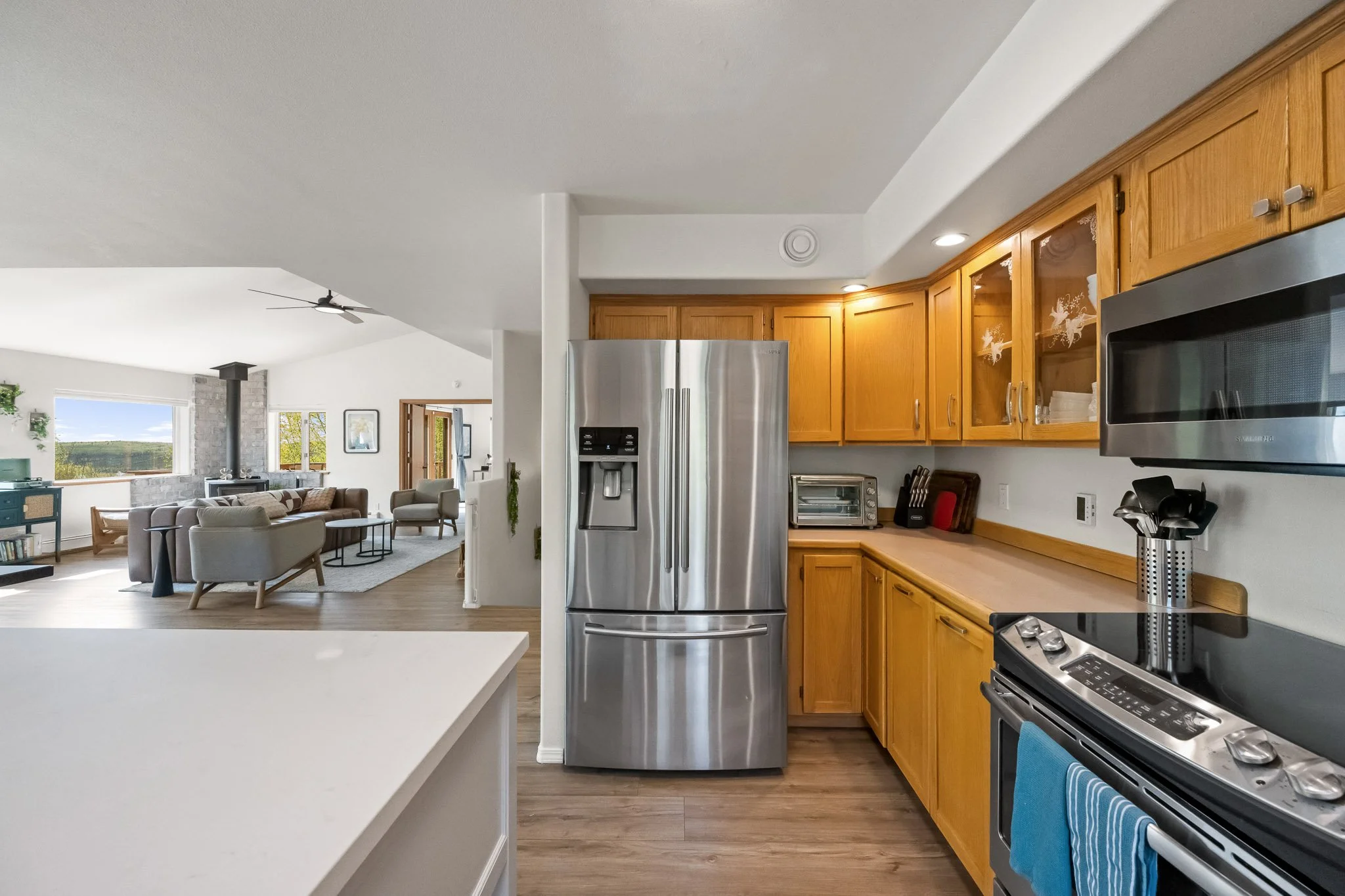 View of a modern kitchen with wooden cabinets, stainless steel refrigerator, and black appliances, opening to a bright living room with large windows and seating area.