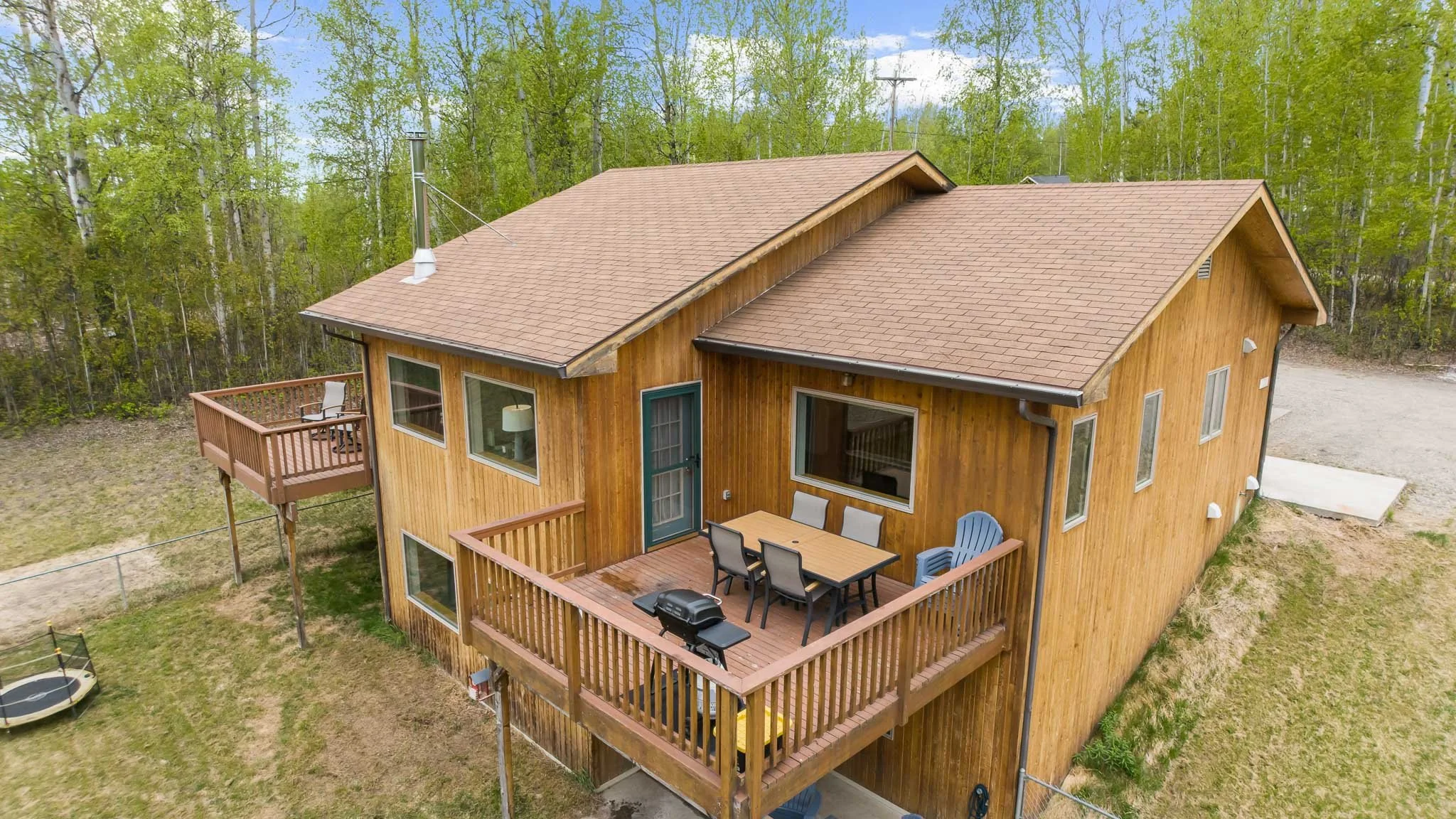 A two-story wooden house with a wrap-around deck, outdoor furniture, and a grill, surrounded by a grass yard and green trees, under a partly cloudy sky.