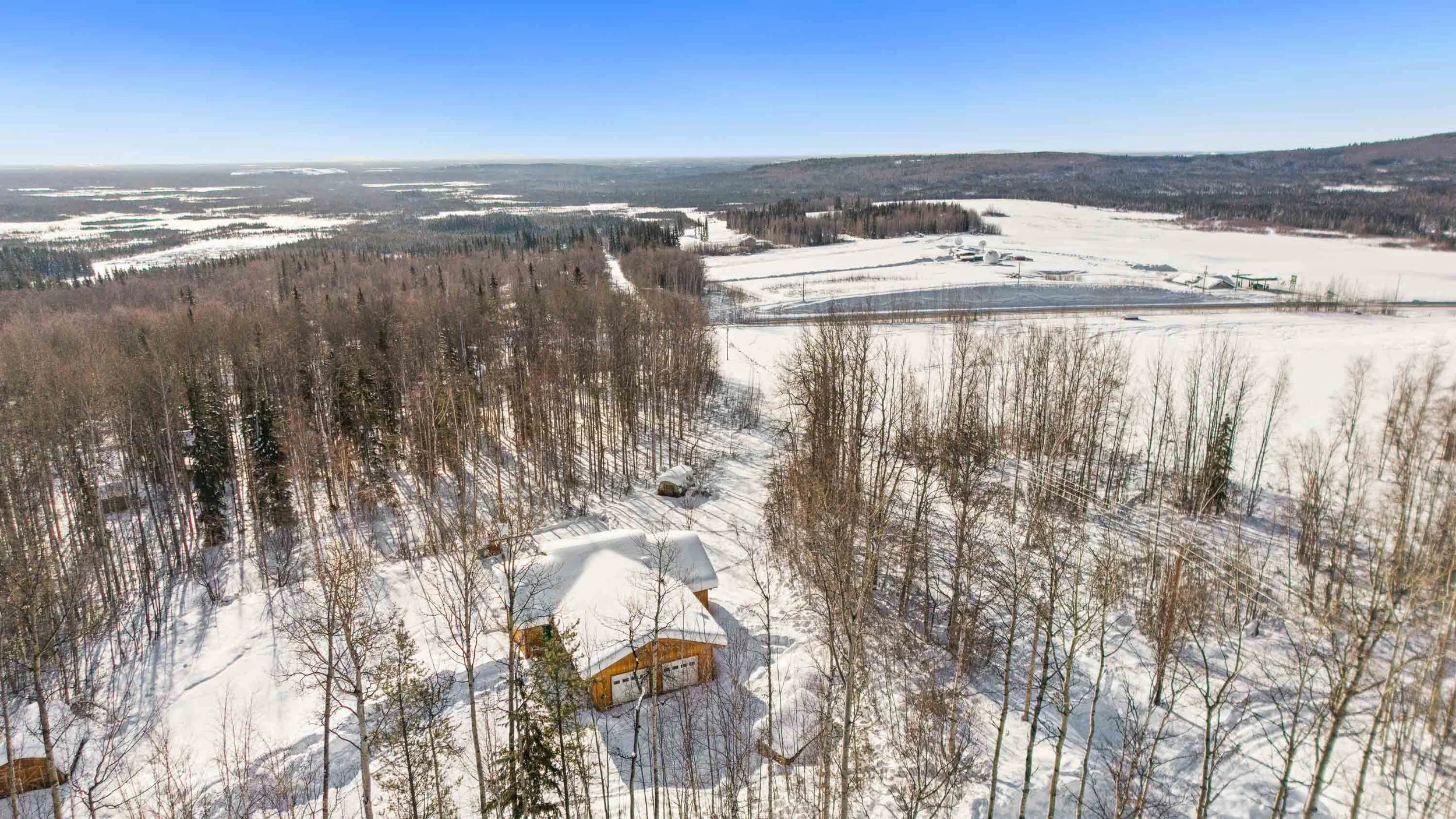 Aerial view of a snowy landscape with trees, open snow-covered fields, and a few houses or cabins, under a clear blue sky.