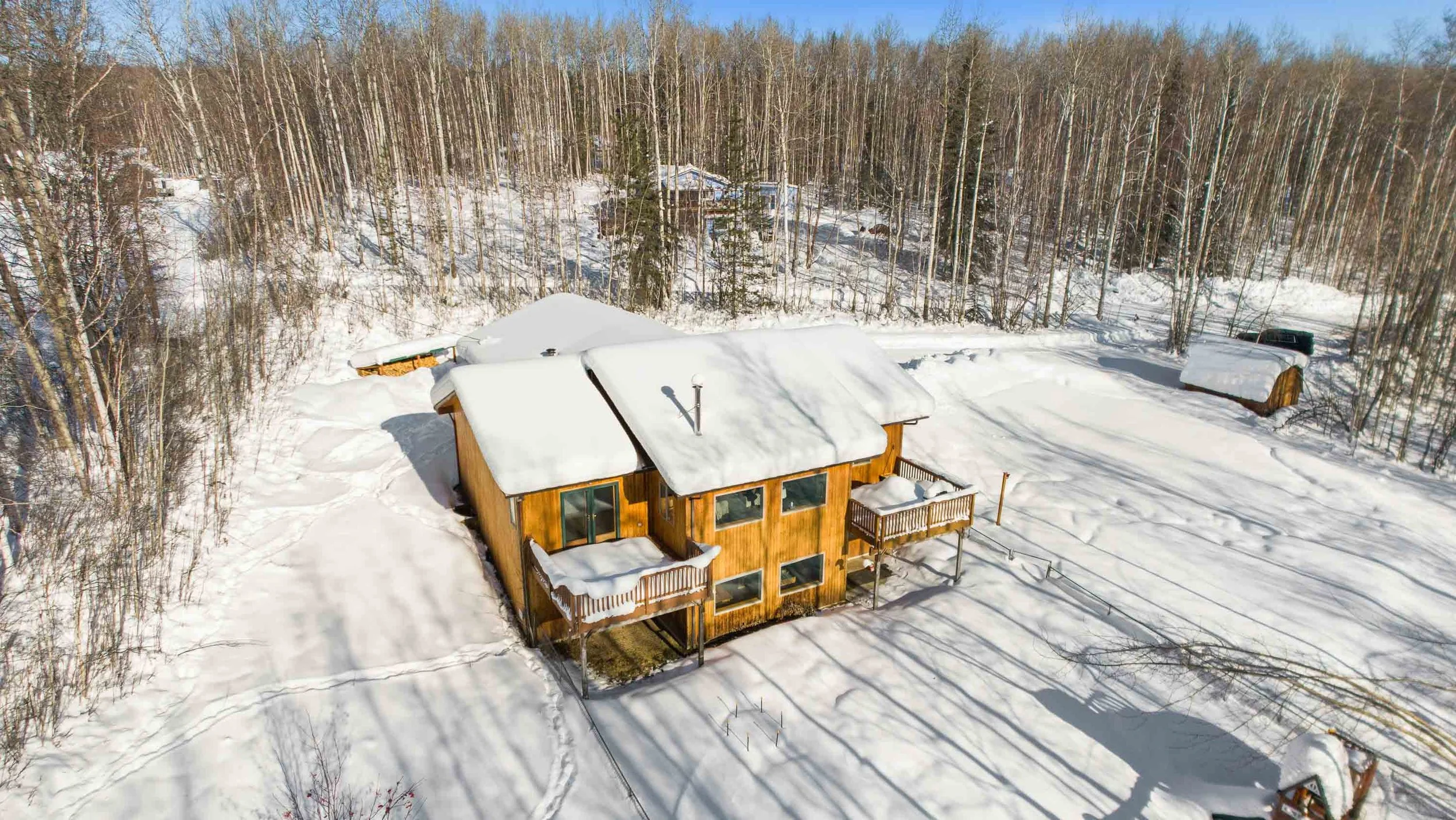 A wooden house covered in snow is surrounded by snow-covered ground and a dense forest of leafless trees. The house has a deck and a chimney, with snow piled on the roof and decking.