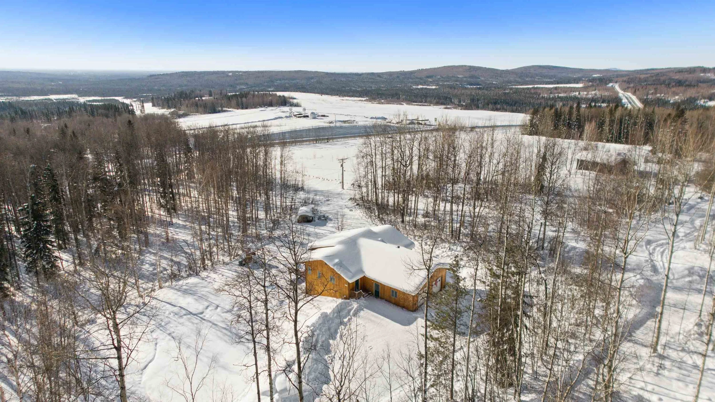An aerial view of a snow-covered landscape with leafless trees, a yellow house with a snow-covered roof, and open fields extending to distant hills under a clear blue sky.