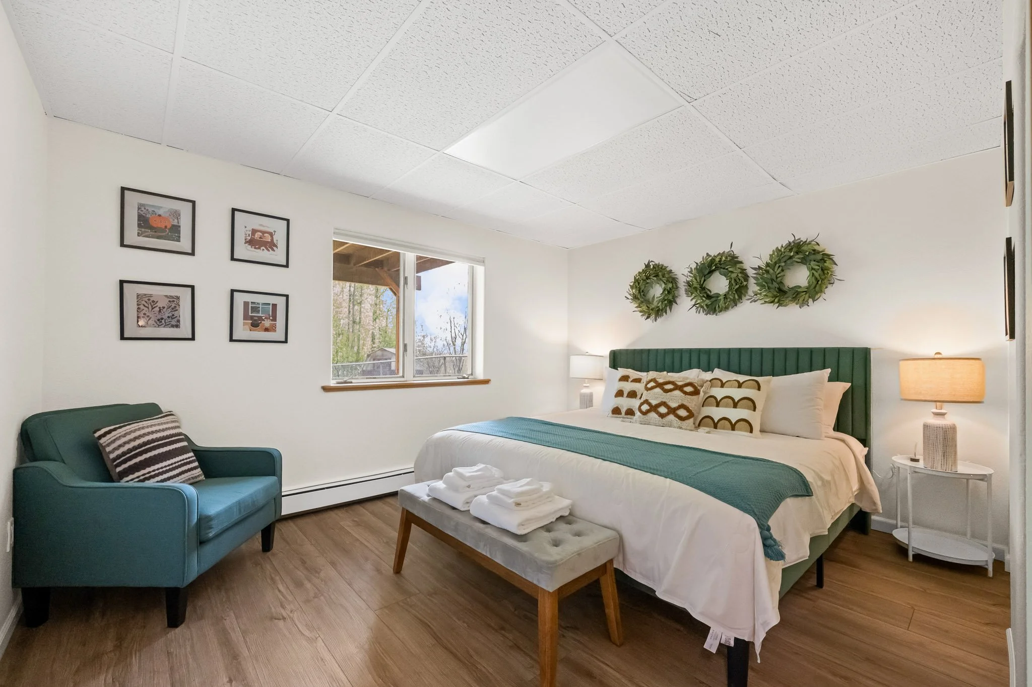 A bedroom with a large bed, green headboard, white bedding, and decorative pillows; side table with a lamp; wall art including framed pictures and green wreaths; window with a view of trees and a deck outside; teal armchair with striped pillow; woode