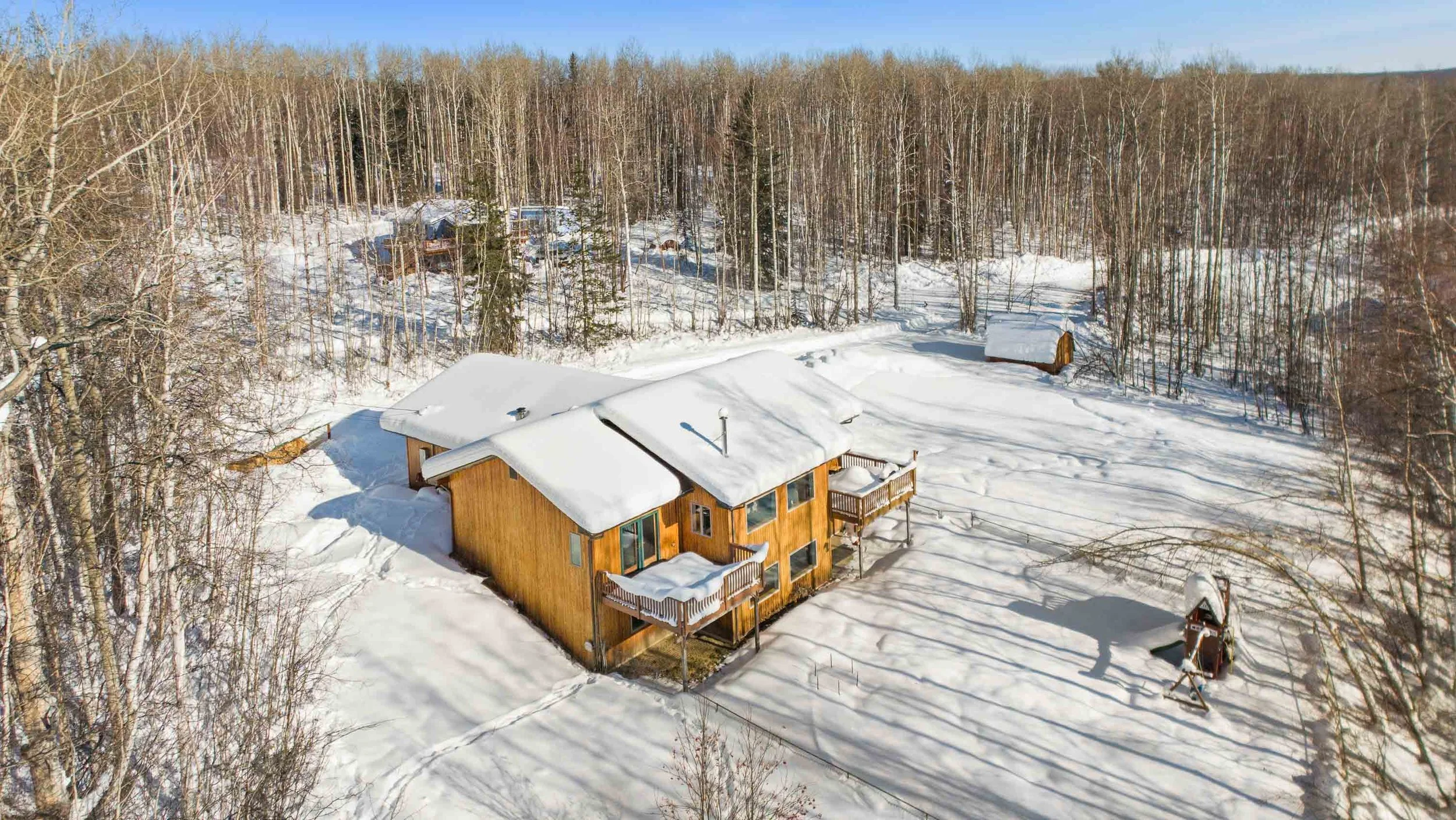 A house covered in snow with a wooden exterior, surrounded by a snowy landscape and a dense forest of leafless trees under a clear blue sky.