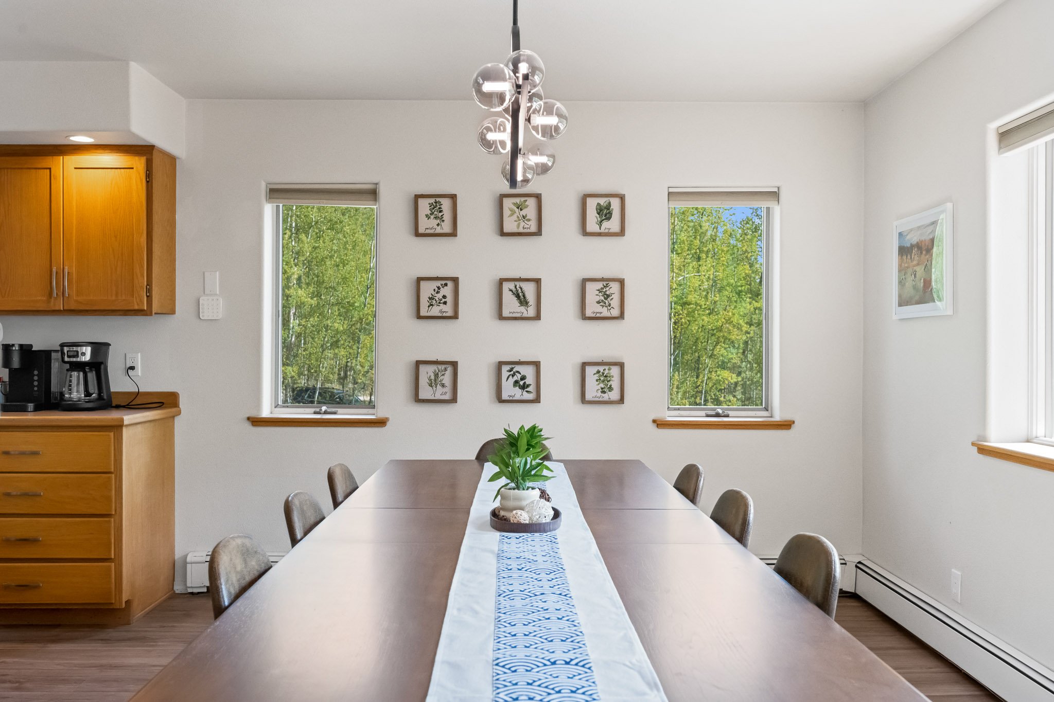 Dining room with a wooden dining table, surrounded by six chairs, a decorative centerpiece, framed botanical prints on white walls, large windows with views of green trees, and a modern chandelier hanging above the table.