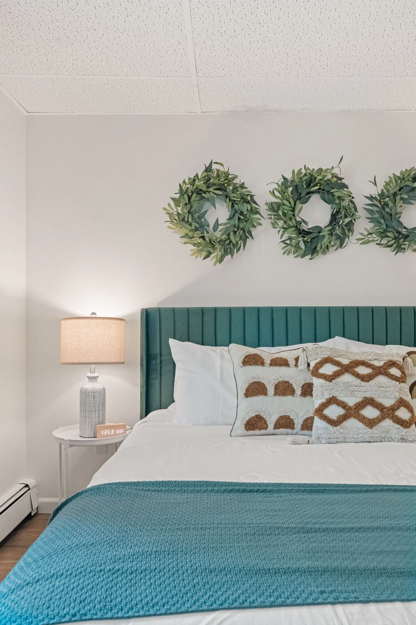 A bedroom with a white brick wall, a green upholstered headboard, white bedding, decorative pillows, a bedside table with a lamp, a digital clock, a turquoise blanket at the foot of the bed, and wall art of greenery wreaths.
