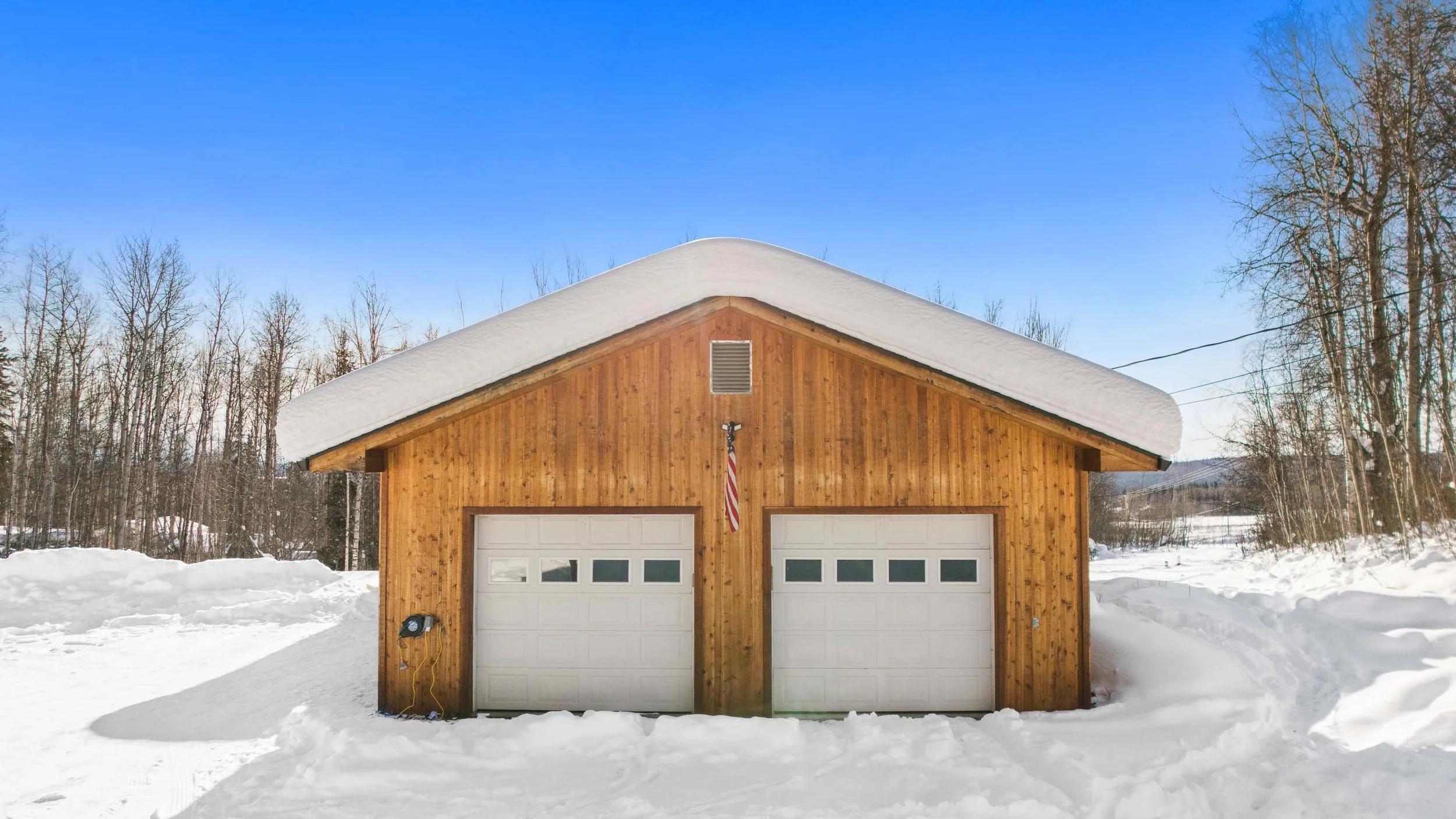 A wooden garage with two white garage doors, snow on the roof and ground, and a wooded background on a clear winter day.