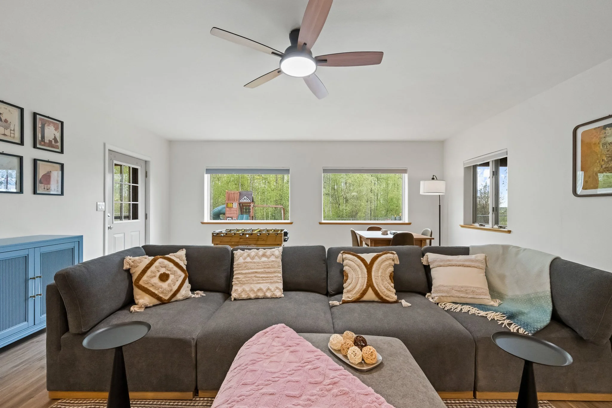 Living room with a large gray sectional sofa with decorative pillows, a pink throw blanket on the ottoman, and two black side tables. Behind the sofa, there is a blue cabinet and framed artwork on the wall, with three windows showing a green outdoor 