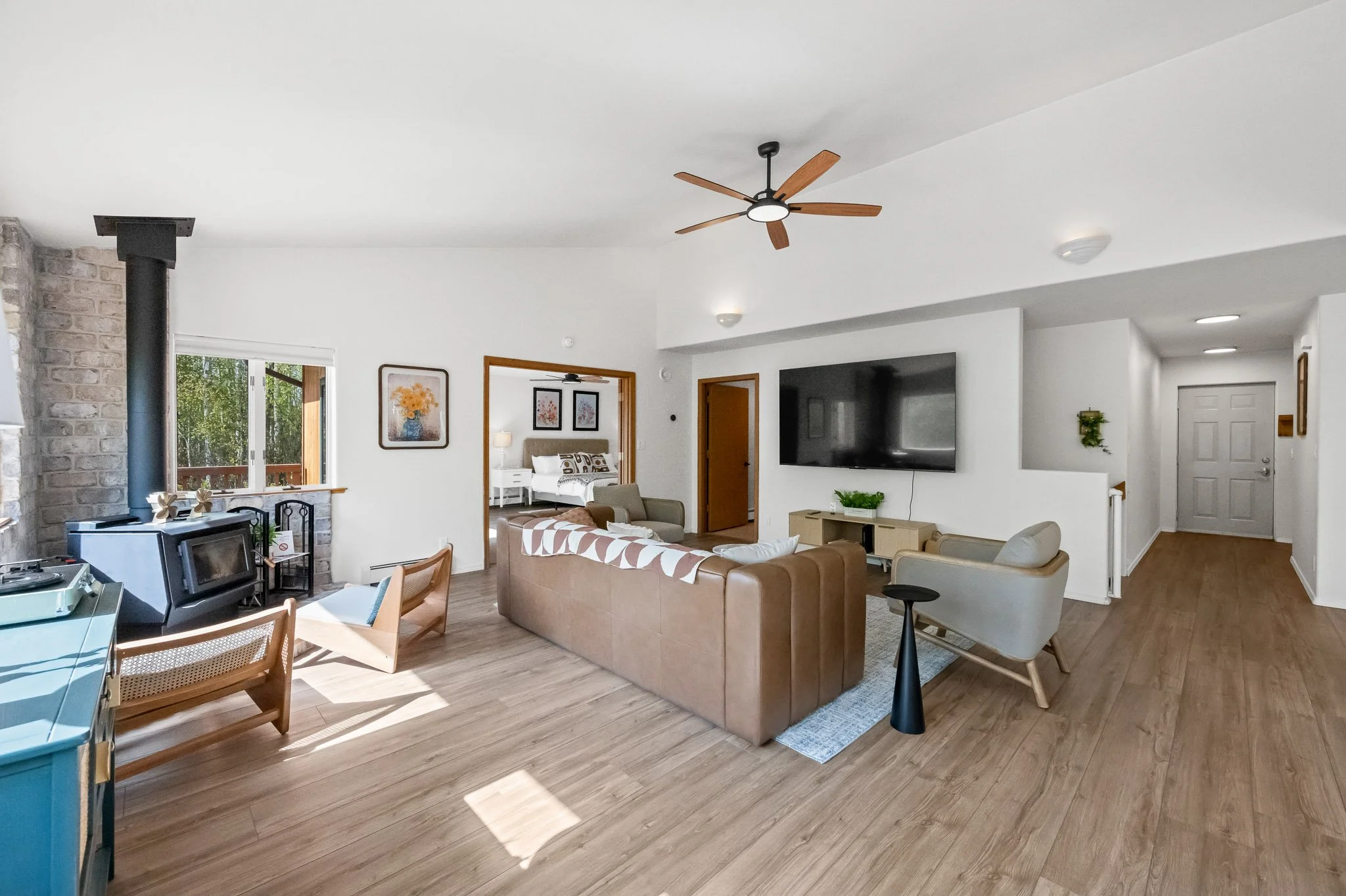 Living room with a beige sectional sofa, a white armchair, a wall-mounted TV, and artwork on the walls. There's a ceiling fan with wooden blades and a window with natural light.