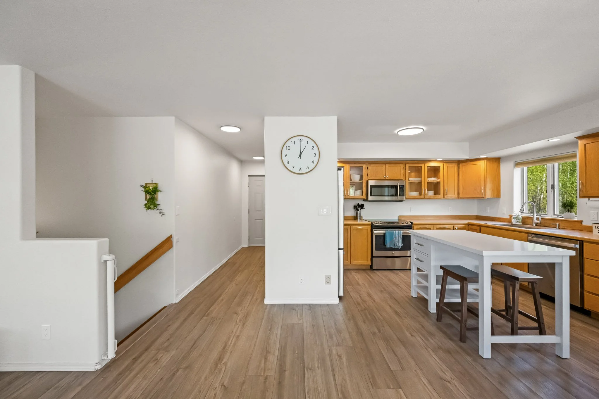 Modern kitchen with wooden cabinets, white island, stainless steel appliances, and large windows revealing greenery outside.