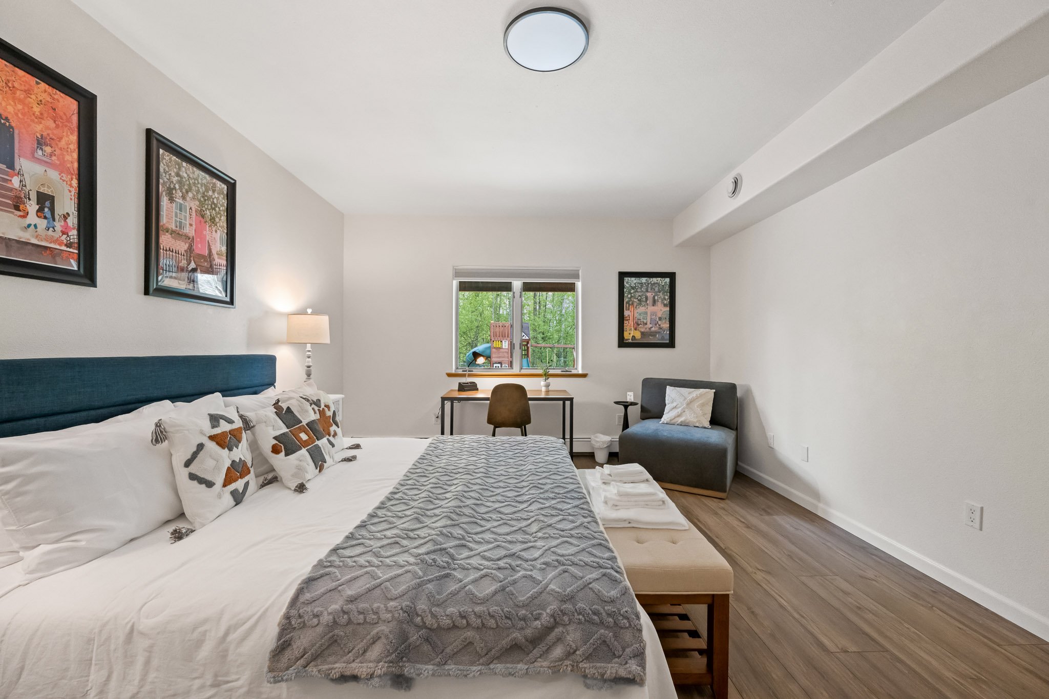 A bedroom with a large bed with pillows, a desk with a chair, a window with a view of trees and a small playhouse outside, and framed pictures on the walls.