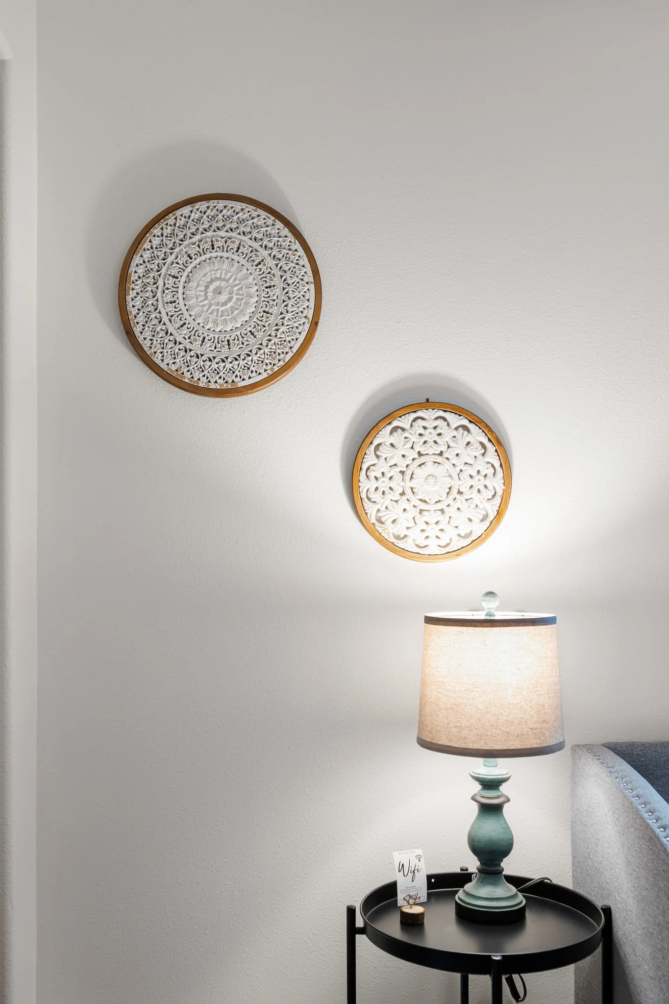 Decorative wall art with intricate circular patterns, a table lamp with a beige shade and light blue base, placed on a black round side table, in a room with white walls and a sofa on the right.