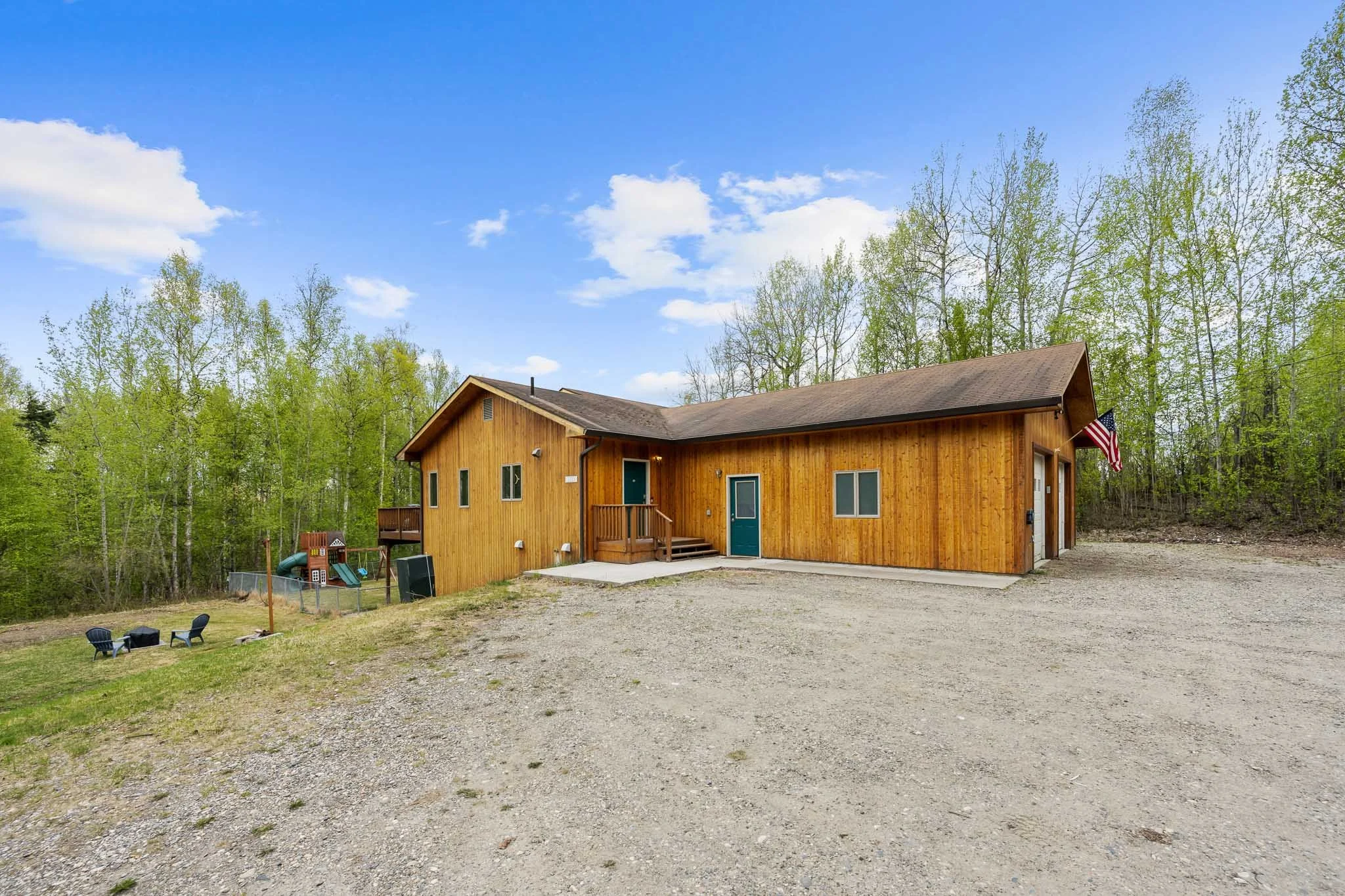 A wooden house with a pitched roof, surrounded by trees with green foliage, on a bright day with a partly cloudy sky. There is a gravel driveway in front of the house, with a small recreational area including a plastic slide and chairs.
