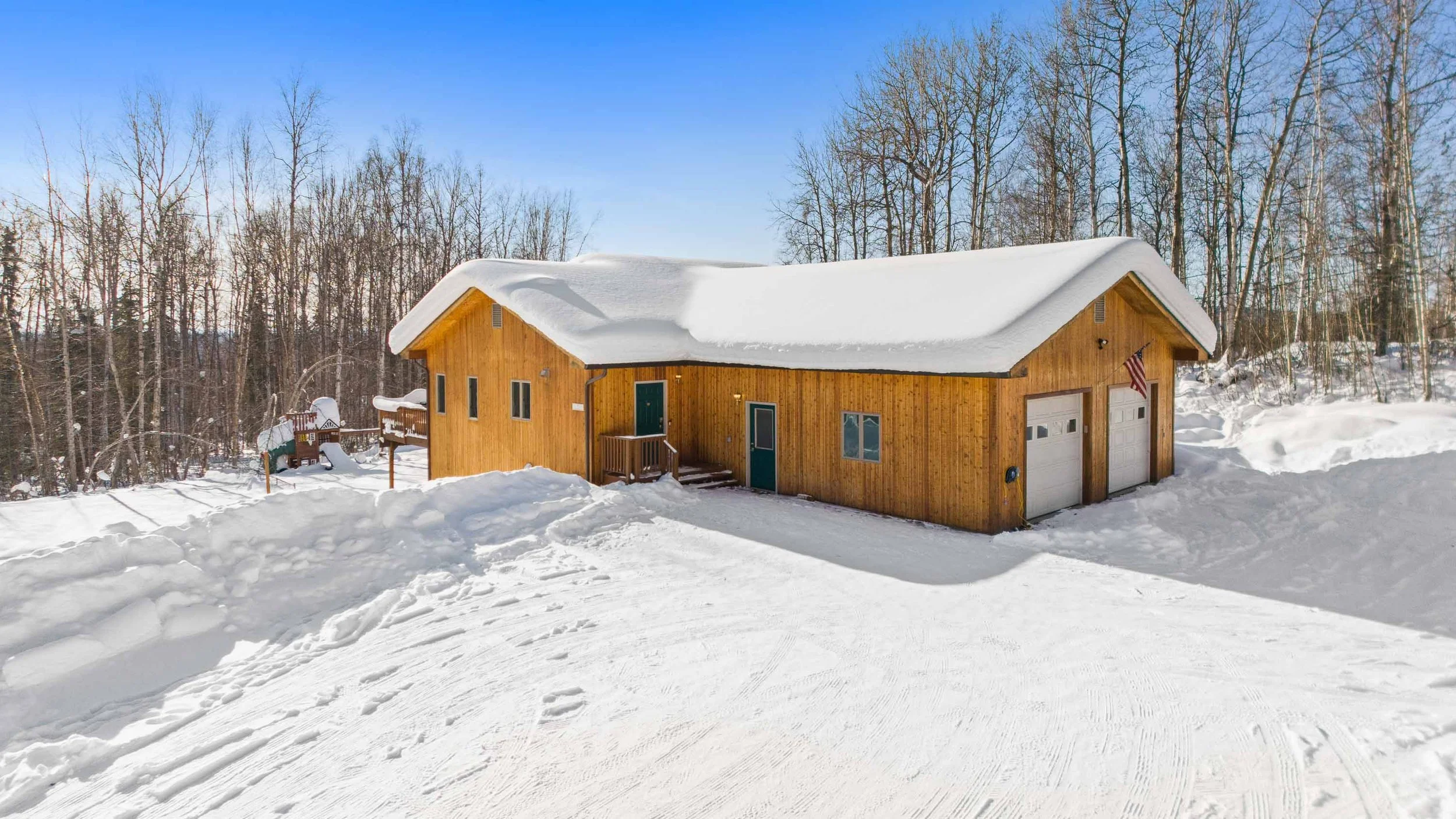 A wooden house covered with snow in a winter landscape, with bare trees in the background and a clear blue sky.