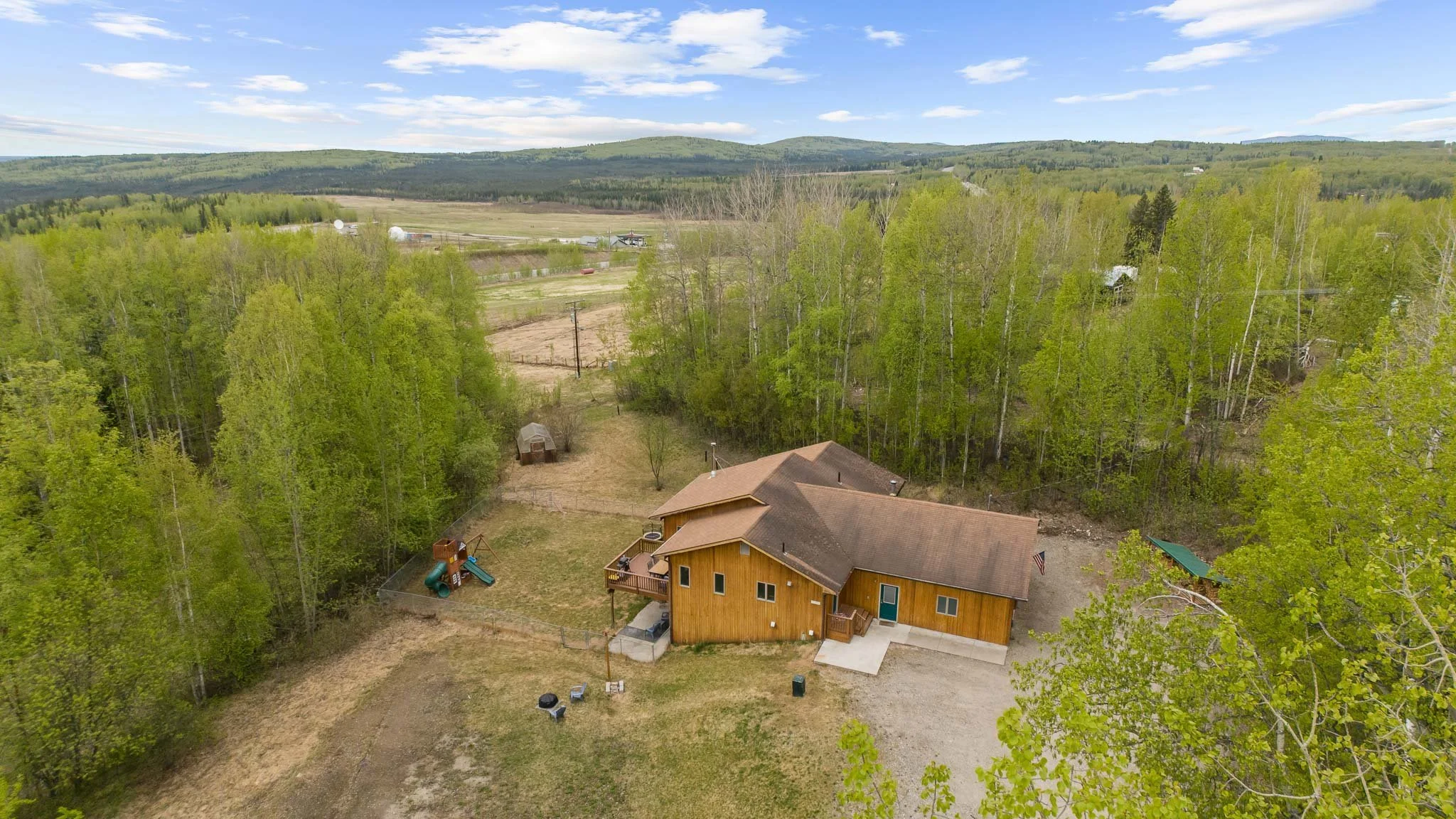 Aerial view of a backyard with a wooden house, a fenced yard, a children's slide and yard toys, surrounded by green trees. Beyond the trees, there are open fields, farms, and hills under a partly cloudy sky.