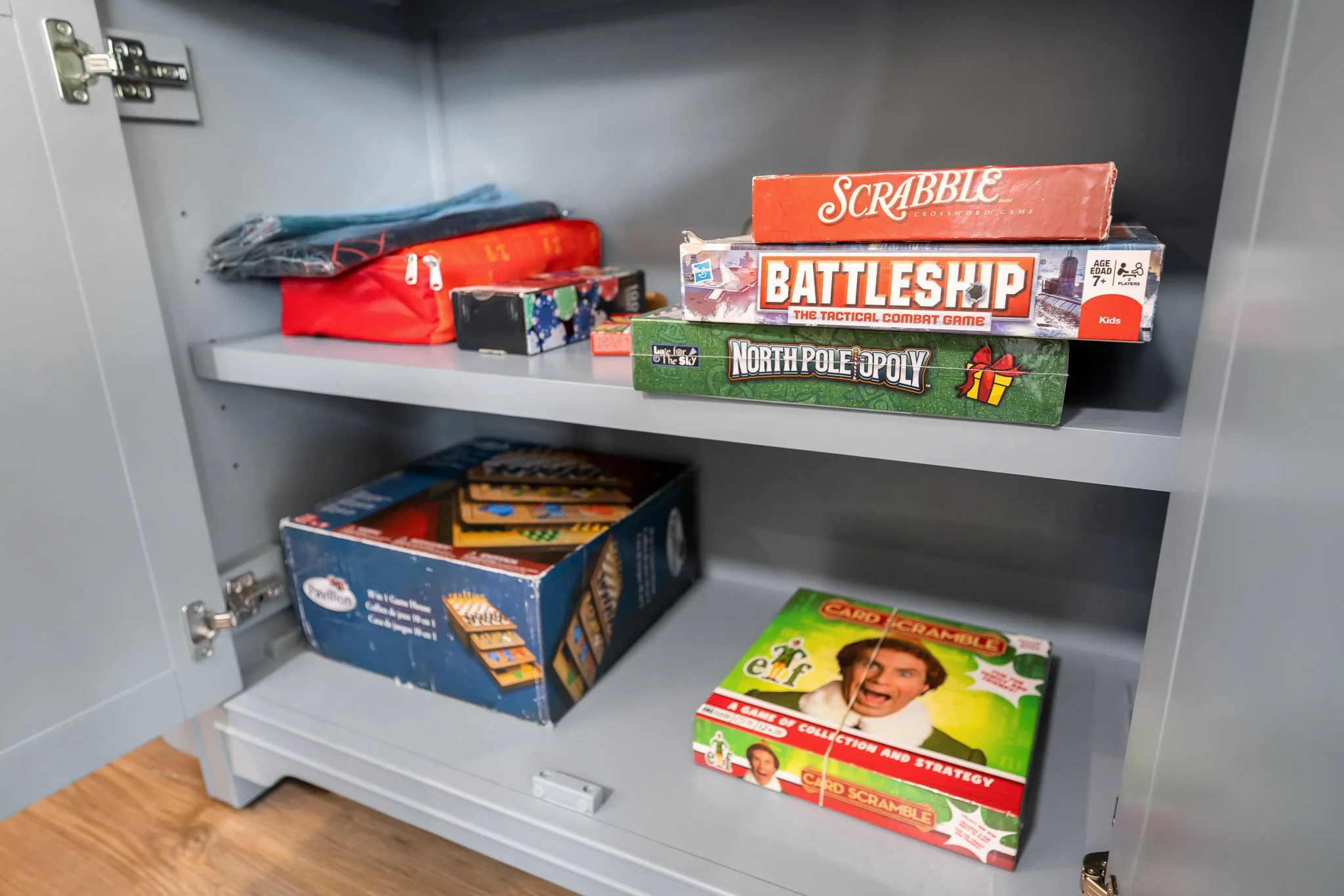 Inside a gray cabinet with various board games on the shelves, including Scrabble, Battleship, North Poleopoly, and a Christmas-themed card game.