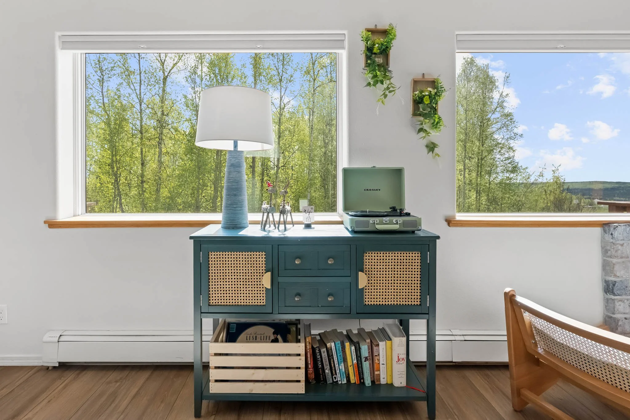 A teal-blue sideboard with rattan cabinet doors, topped with a white lamp, decorative metallic reindeer figures, and a small sign, set against large windows showcasing green trees and a blue sky.