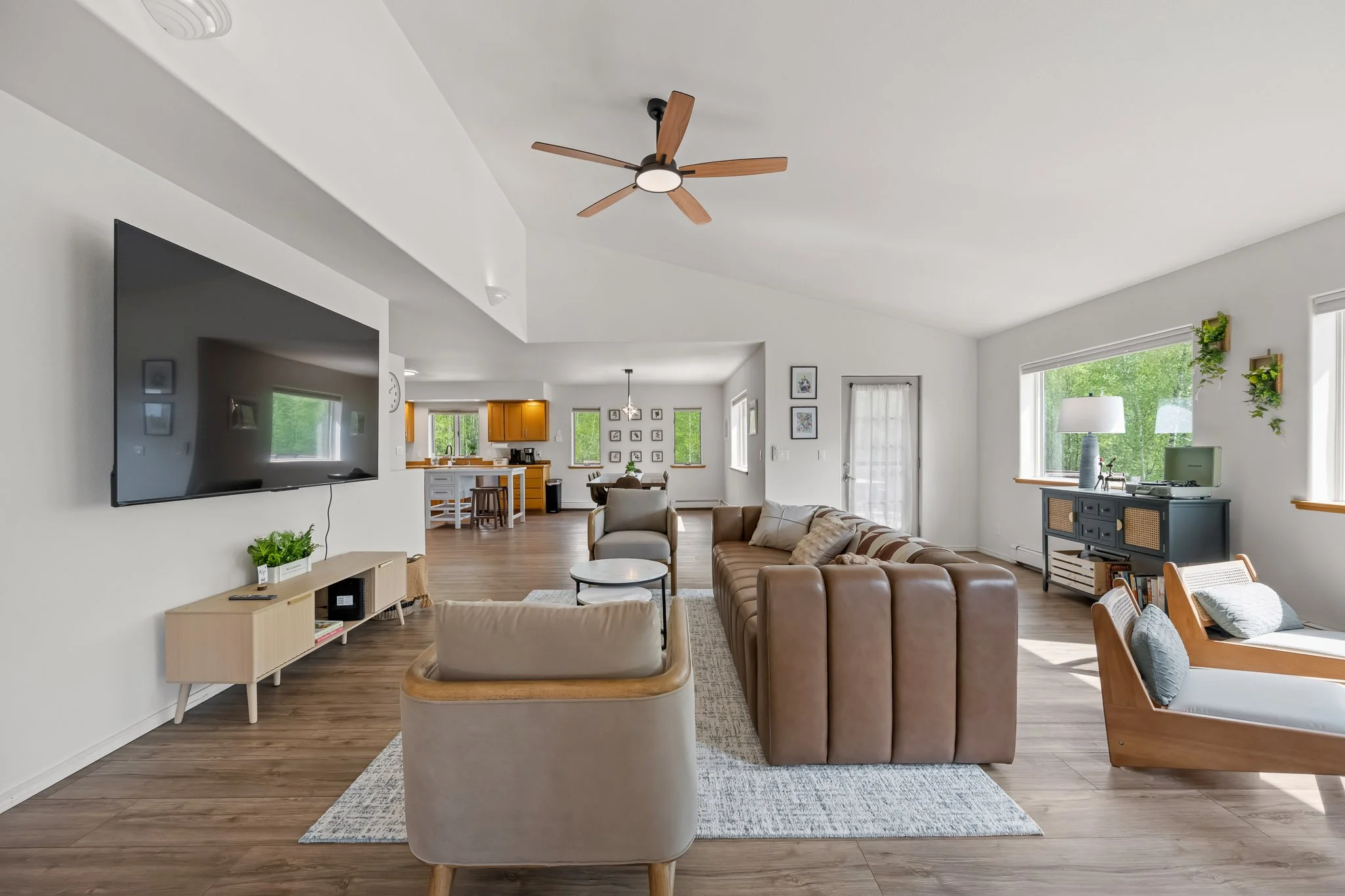 Spacious living room with a ceiling fan, large windows letting in natural light, a brown leather sofa, a beige armchair, a white coffee table, a TV mounted on the wall, and a view of the dining and kitchen area in the background.