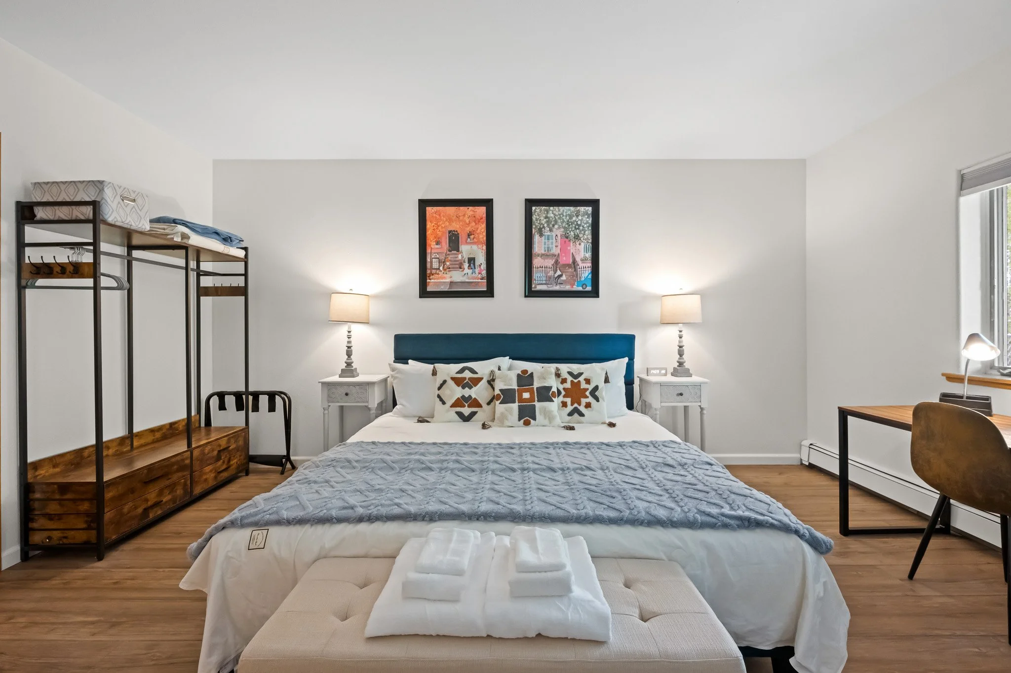 A neatly made bed with white sheets, a blue headboard, and decorative pillows, situated between two white nightstands with lamps. The room features framed artwork on the wall, a wooden dresser on the left, and a small table and chair on the right.