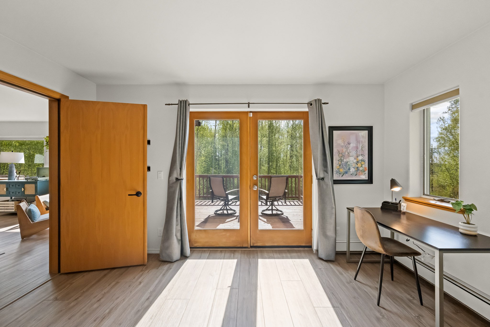 Living room with wooden sliding doors leading to a balcony with outdoor chairs, and a view of green trees outside. There's a desk with a lamp and plant near the window, and framed artwork on the wall.