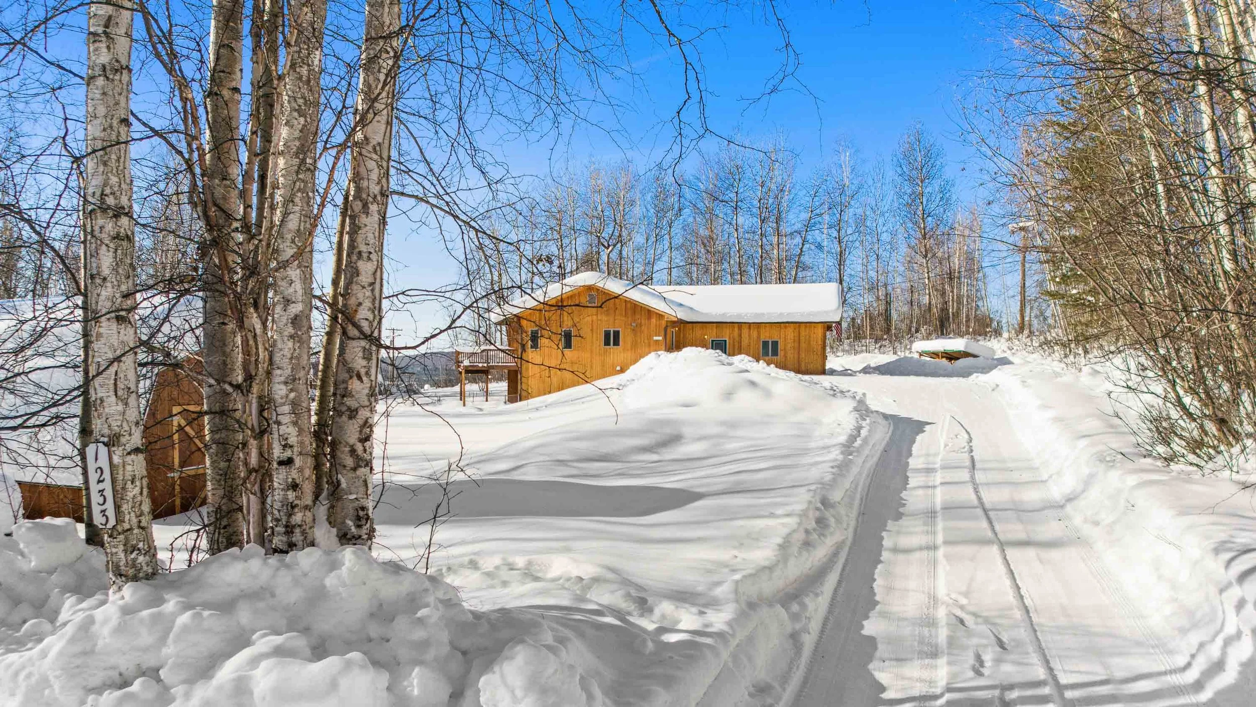 A snowy landscape with a wooden house and surrounding trees under a clear blue sky, with snow-covered ground and tire tracks in the snow.