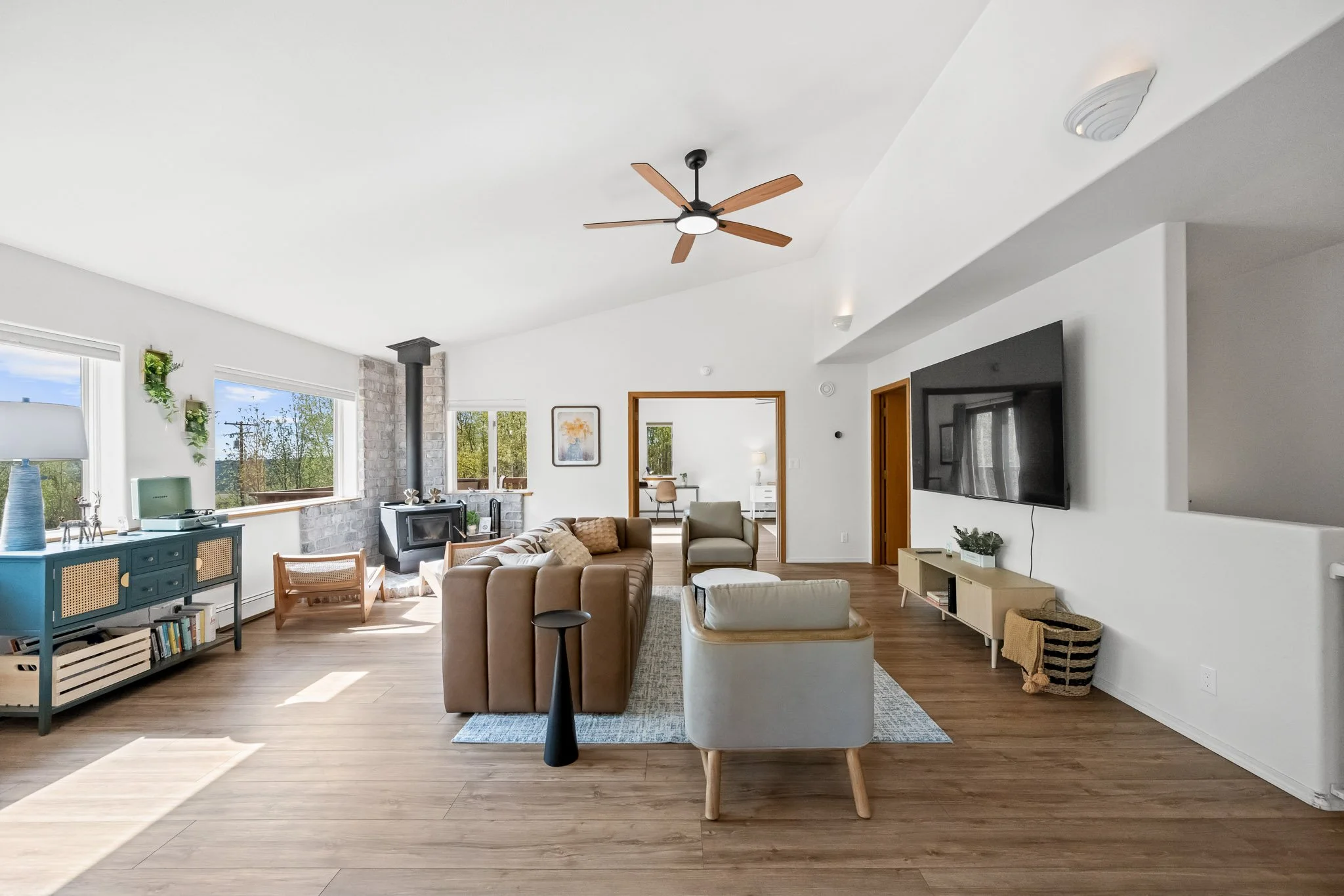 Living room with white walls, wooden flooring, a ceiling fan, large windows, a combination of beige and white furniture, a wall-mounted TV, and decorative plants.