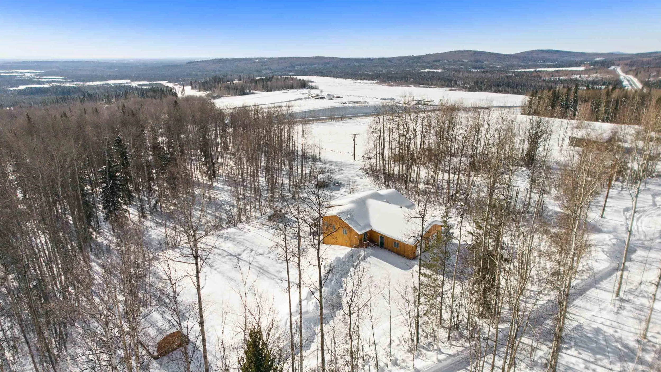 A snow-covered rural landscape featuring a wooden house, leafless trees, fields, and distant hills under a clear blue sky.