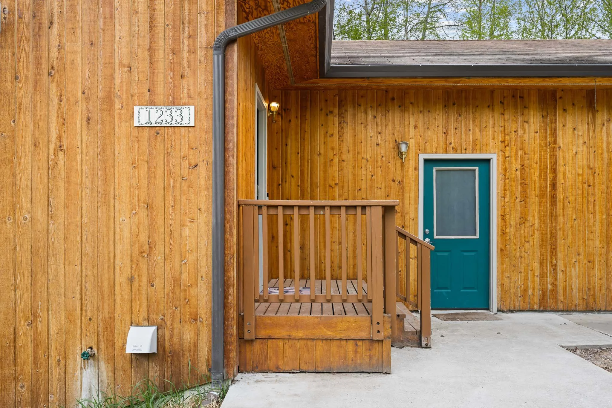 View of a house exterior with vertical wooden siding, a teal door, small porch with wooden railing, and house number 1233 on a white plaque. Part of the roof and a blue sky with green trees are visible.
