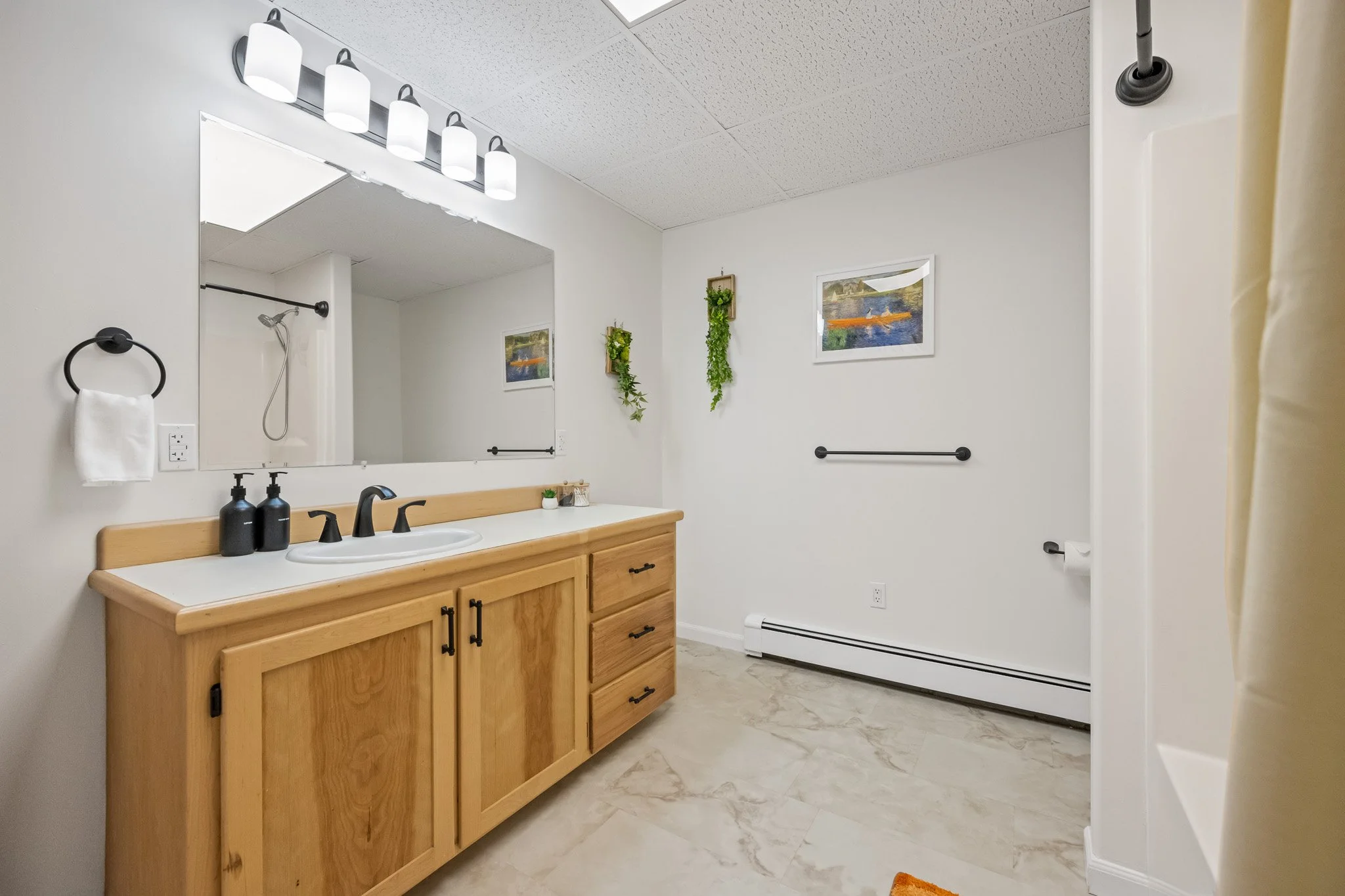 Bathroom with wooden vanity, white countertop, black faucet, large mirror, wall-mounted light fixture, small framed paintings, and hanging plants on the wall.