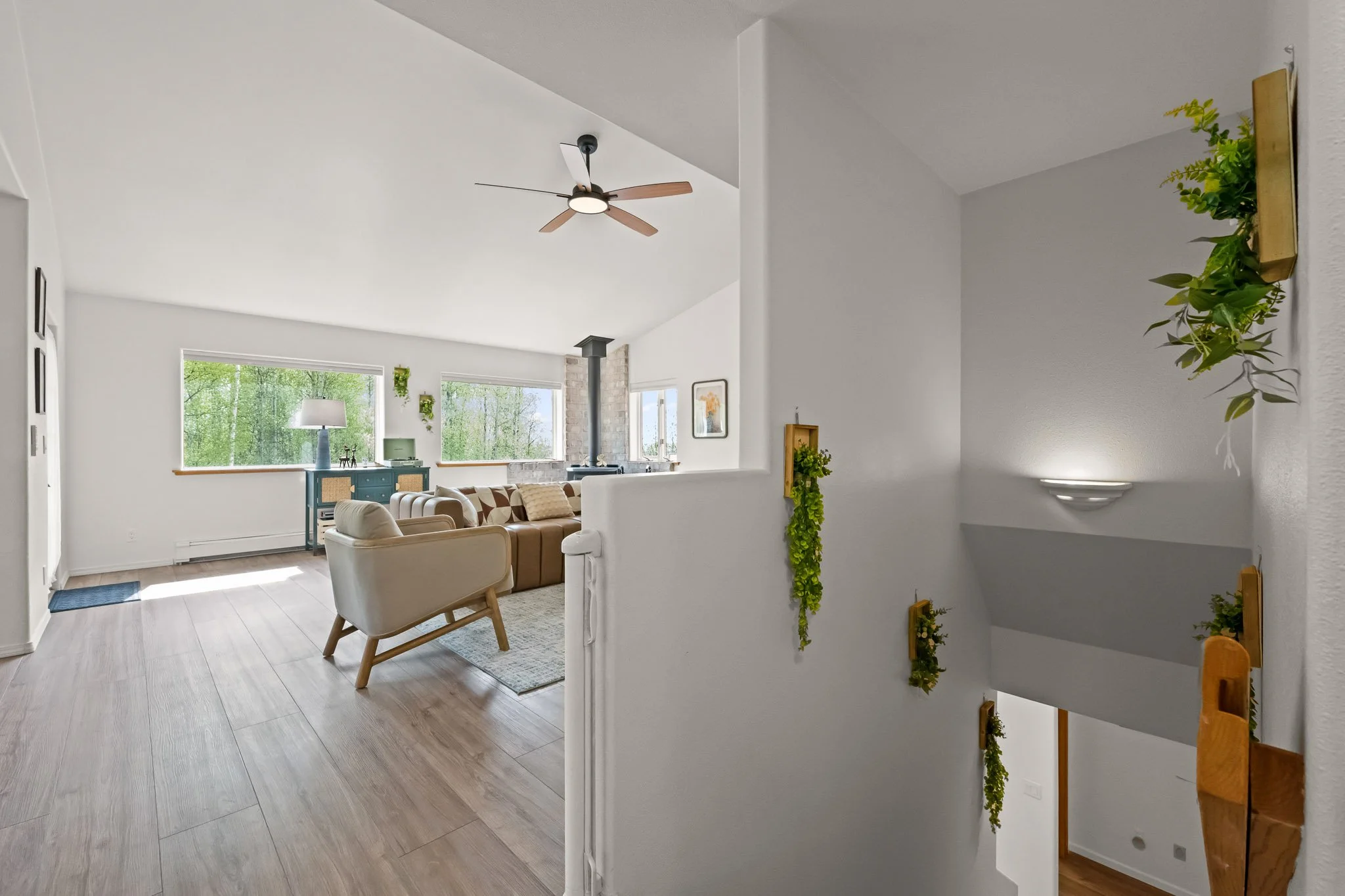 Living room with large windows, a ceiling fan, beige sofa, blue cabinet, and wall decor with indoor plants.