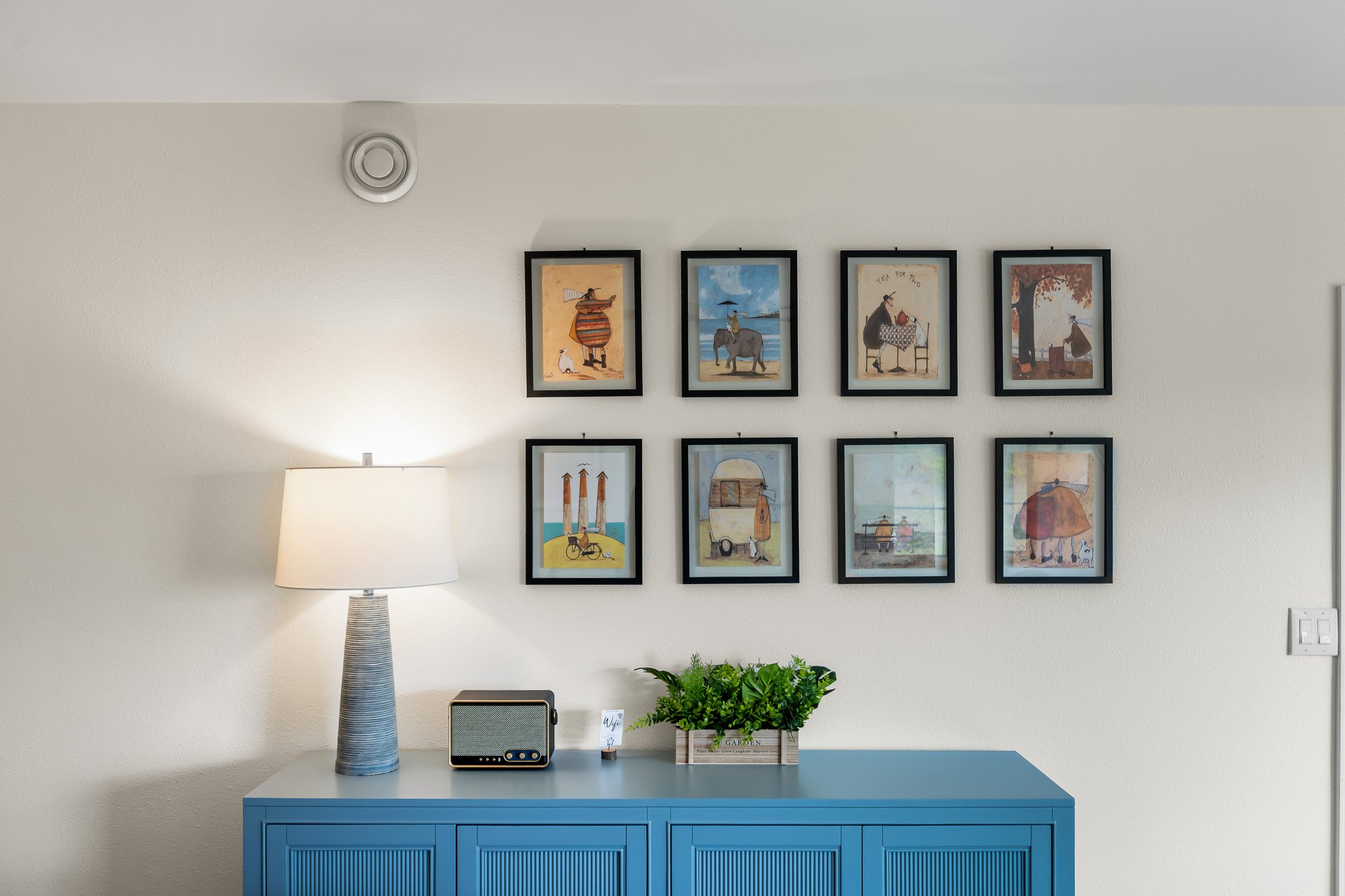 A wall decorated with a series of framed artwork featuring whimsical scenes with animals. Below the artwork, a blue cabinet holds a lamp, a small speaker, and a plant in a wooden crate.