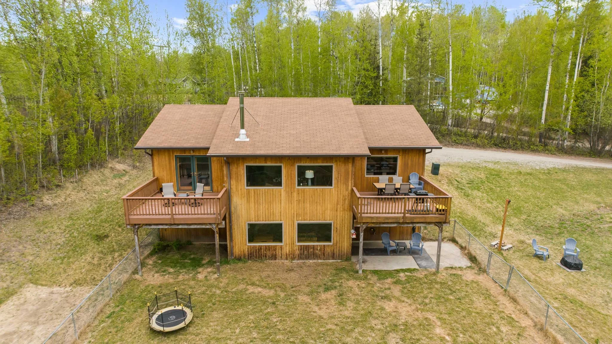 An aerial view of a two-story wooden house with a brown shingled roof, surrounded by a grassy yard and dense green trees. The house has two balconies with outdoor furniture, and a small trampoline is in the yard.