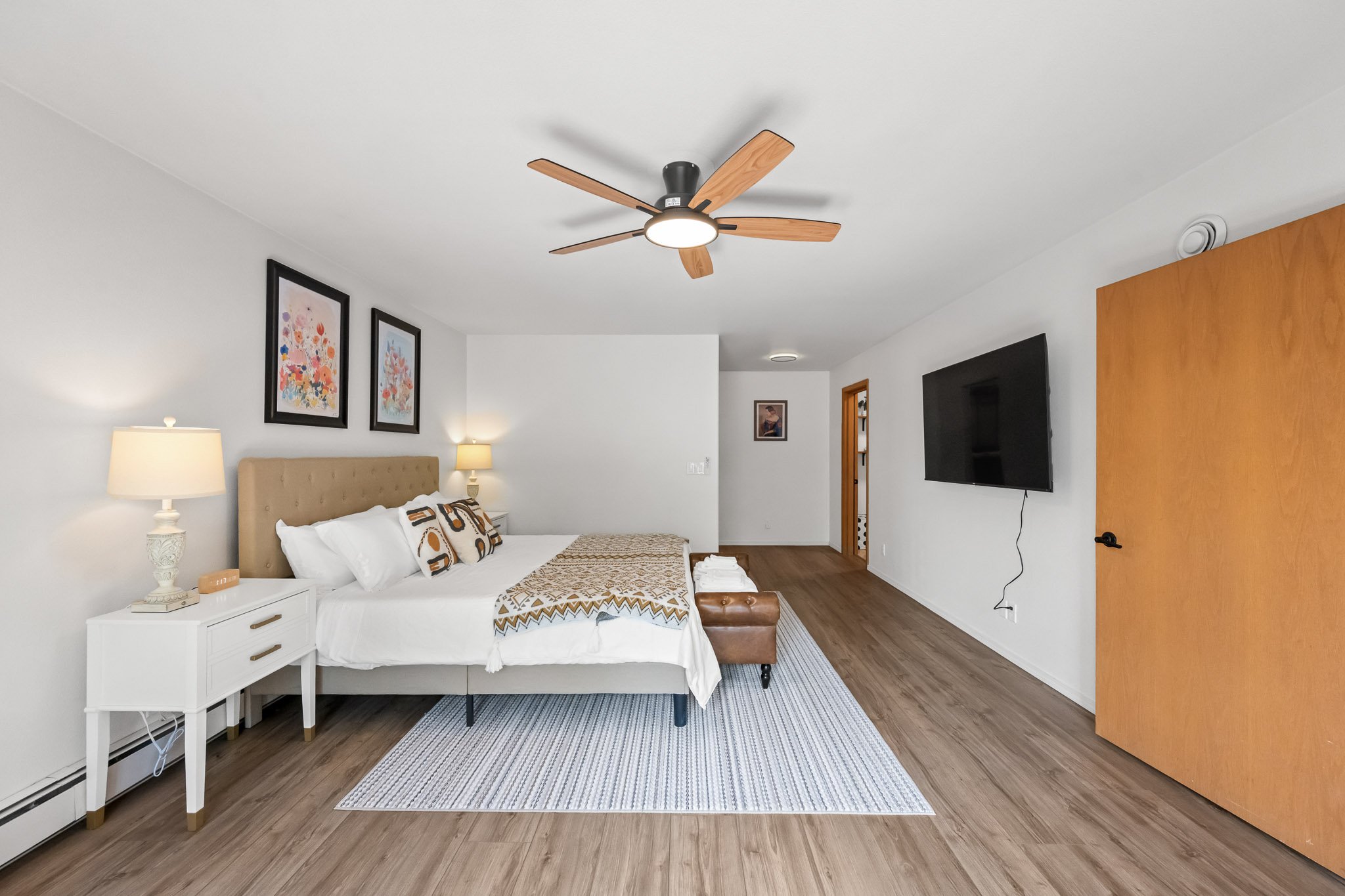 A bedroom with a bed, white nightstand, table lamps, framed floral art, a flat-screen TV, a ceiling fan, and wooden flooring.