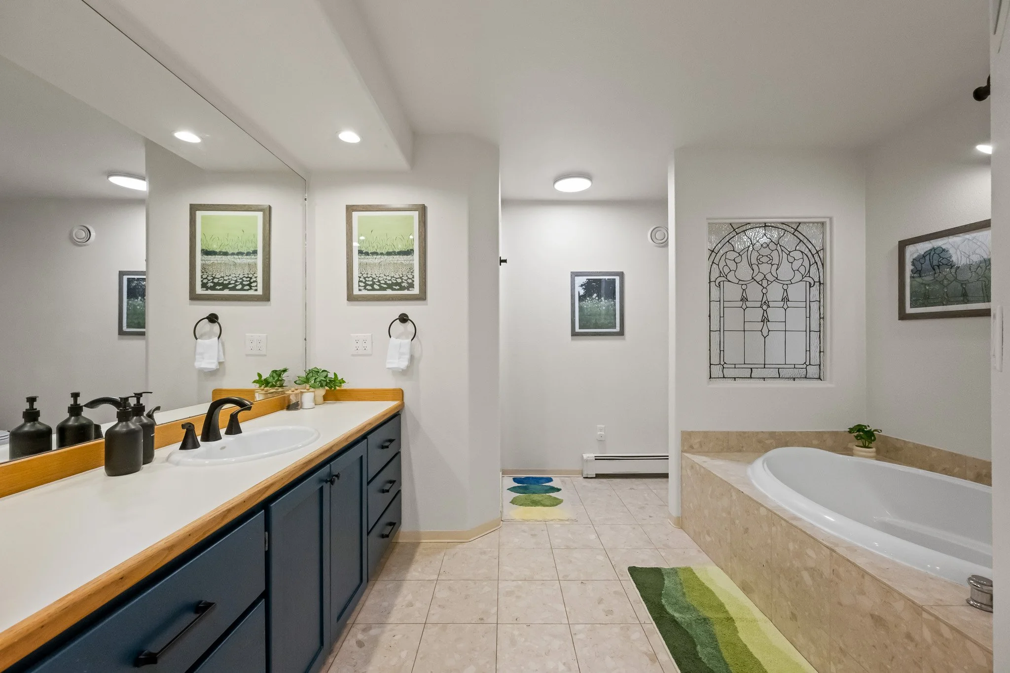 A bathroom with a large mirror above a blue vanity with black soap dispensers, framed pictures of green fields, a towel ring with a white towel, a small potted plant, and a double sink. There is a window with stained glass, a bathtub with a beige sur