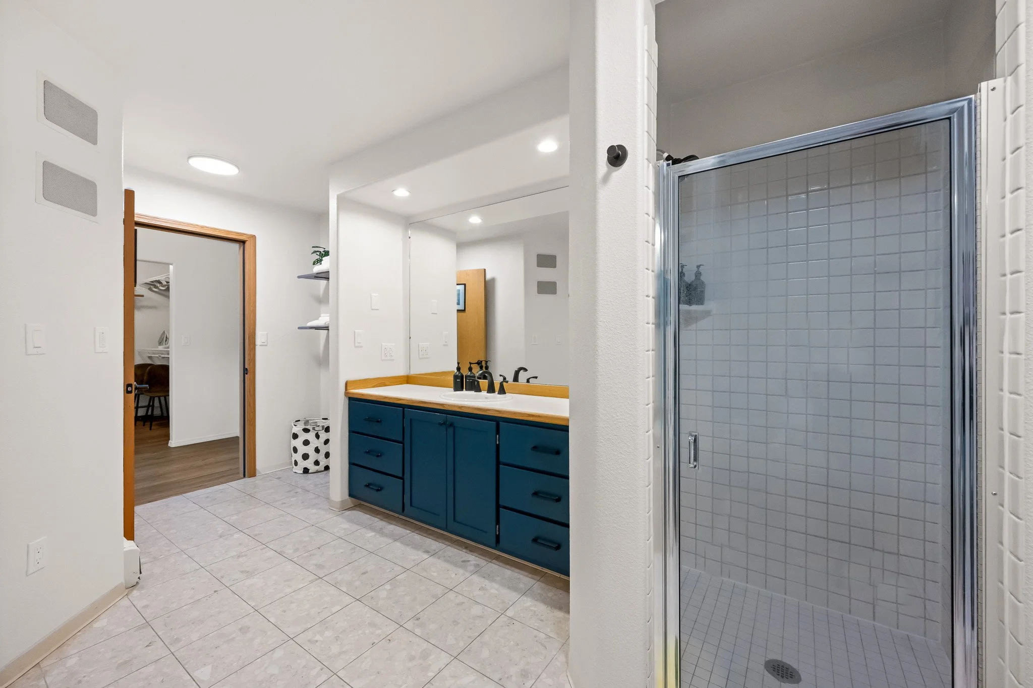 Bathroom with tiled floor, a blue vanity with a white countertop, a mirror, a shower with white tiled walls, and a doorway leading to a walk-in closet.
