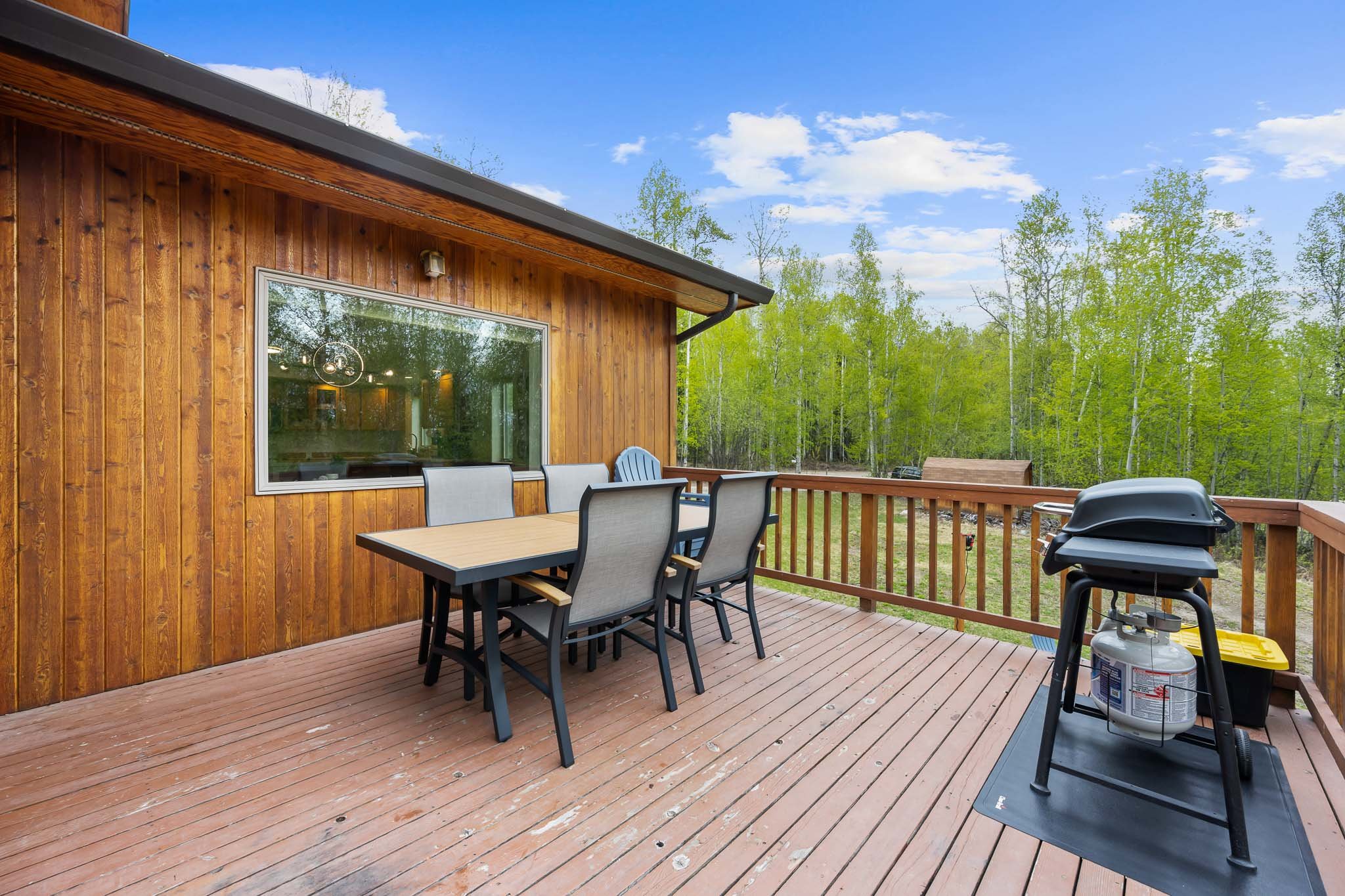 Wooden deck with outdoor dining table and chairs, and a propane grill, overlooking a green forest under a blue sky with clouds.