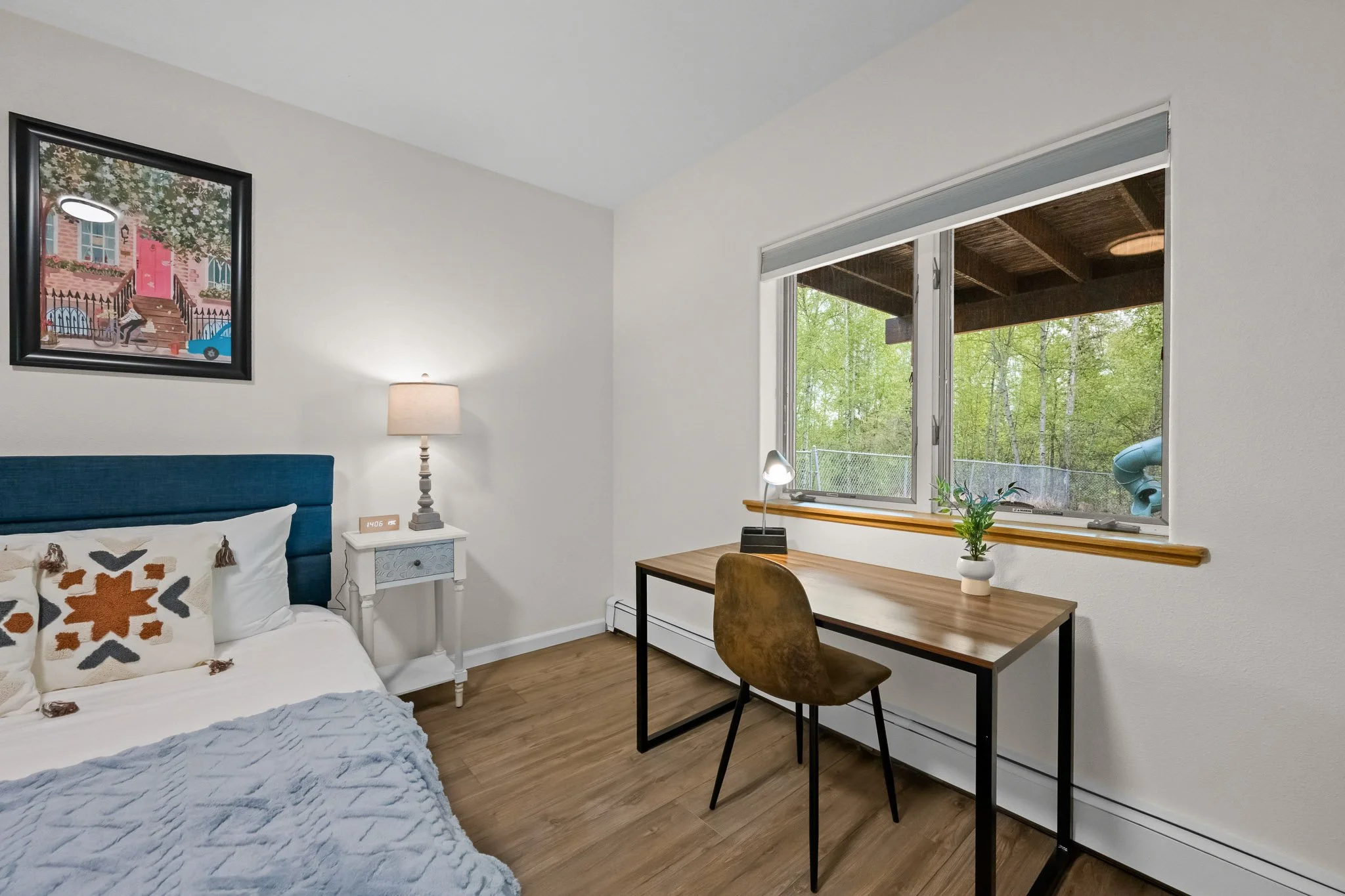 A cozy bedroom with a bed that has a white blanket and decorative pillows, a nightstand with a lamp, a window with a view of green trees, a desk with a chair, a potted plant, and a framed artwork on the wall.