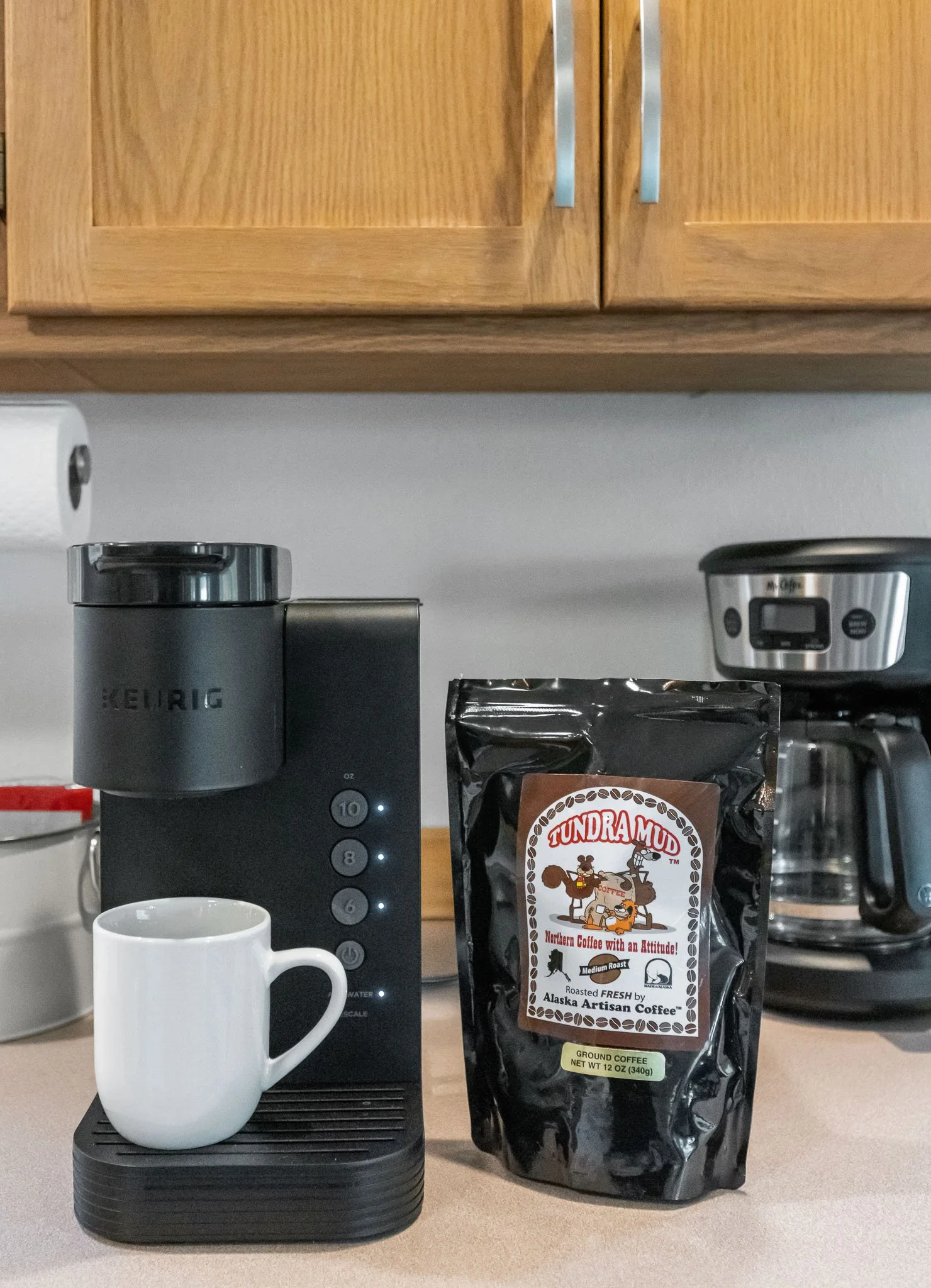 A coffee setup with a Keurig coffee machine, a white coffee mug, and a bag of Tundra Mud coffee on a kitchen counter.