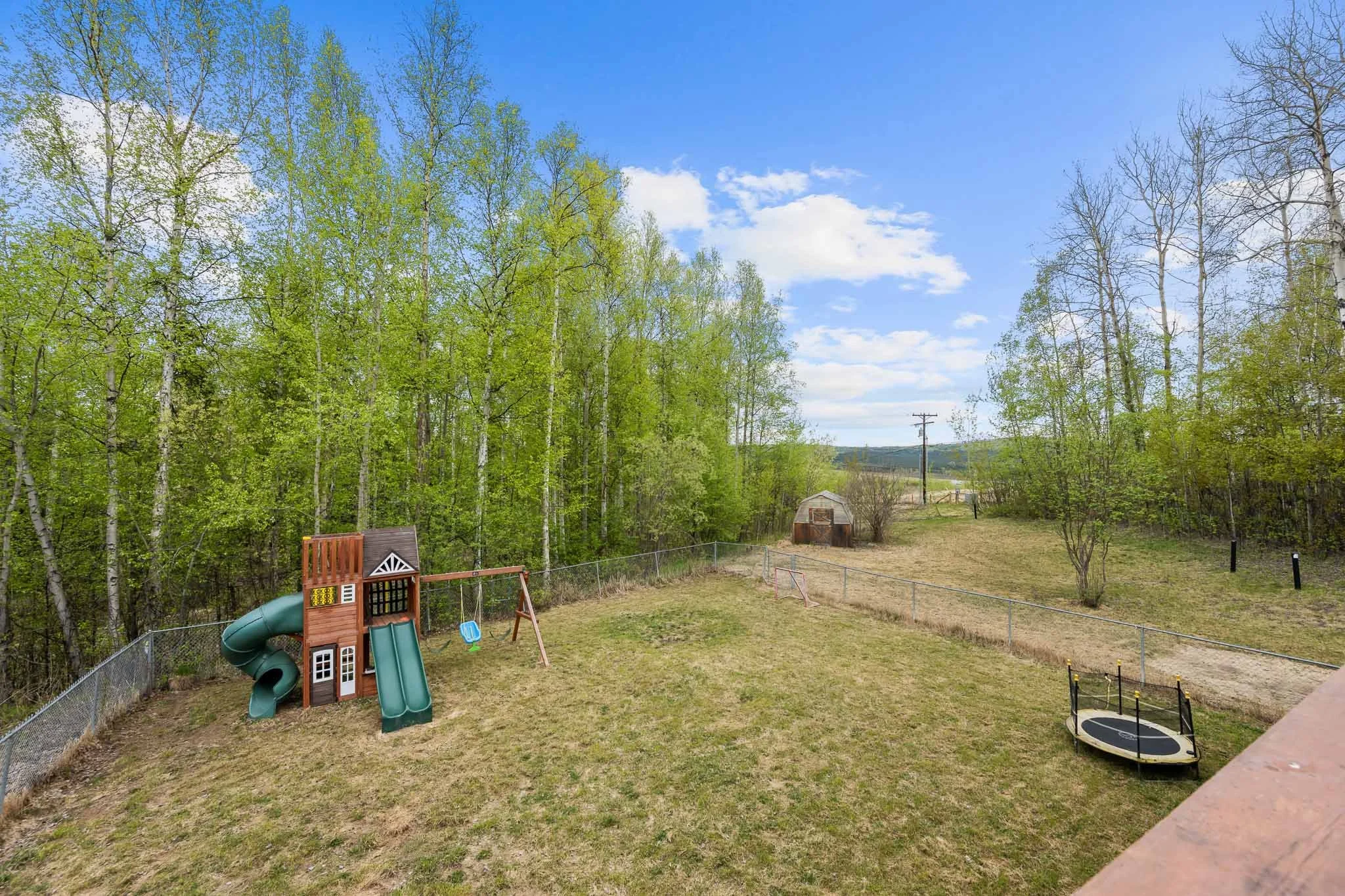 Backyard with playset, trampoline, trees, and a shed under a blue sky with clouds.