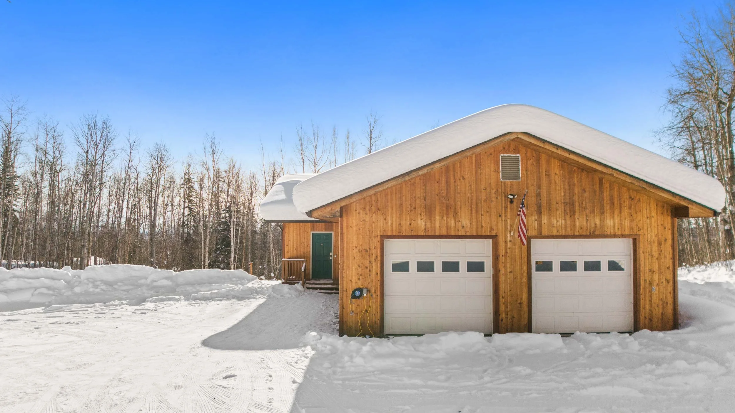 A wooden garage with a snow-covered roof, two white garage doors, and a green front door. An American flag is hanging beside the garage. Snow covers the driveway and ground, with a backdrop of leafless trees under a clear blue sky.