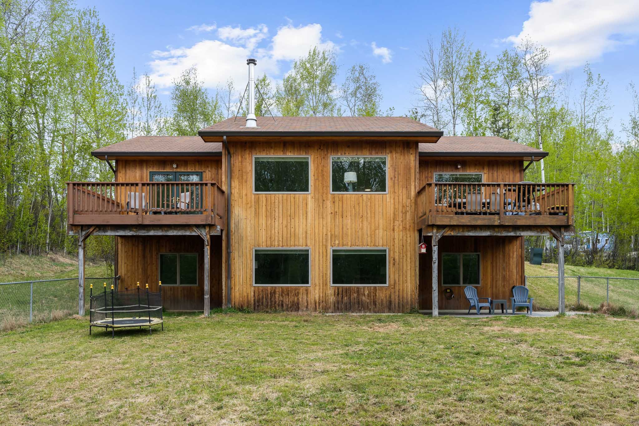 Back view of a two-story wooden house with a balcony on the upper level, large windows, and outdoor furniture, situated in a grassy yard with a chain-link fence, surrounded by trees and under a partly cloudy sky.