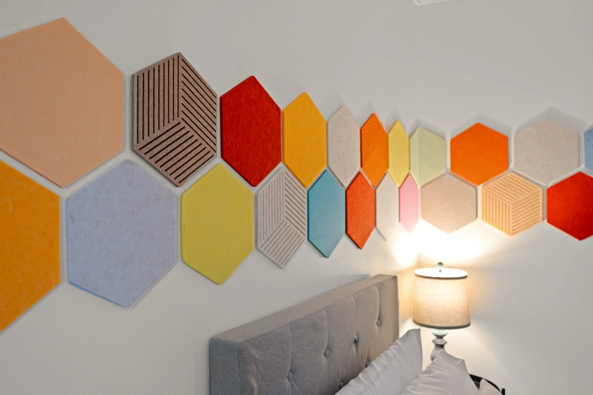 A decorative wall with colorful hexagonal tiles in shades of orange, yellow, blue, white, beige, and brown arranged in a pattern, next to a gray upholstered bench and a table lamp with a beige shade and white base.