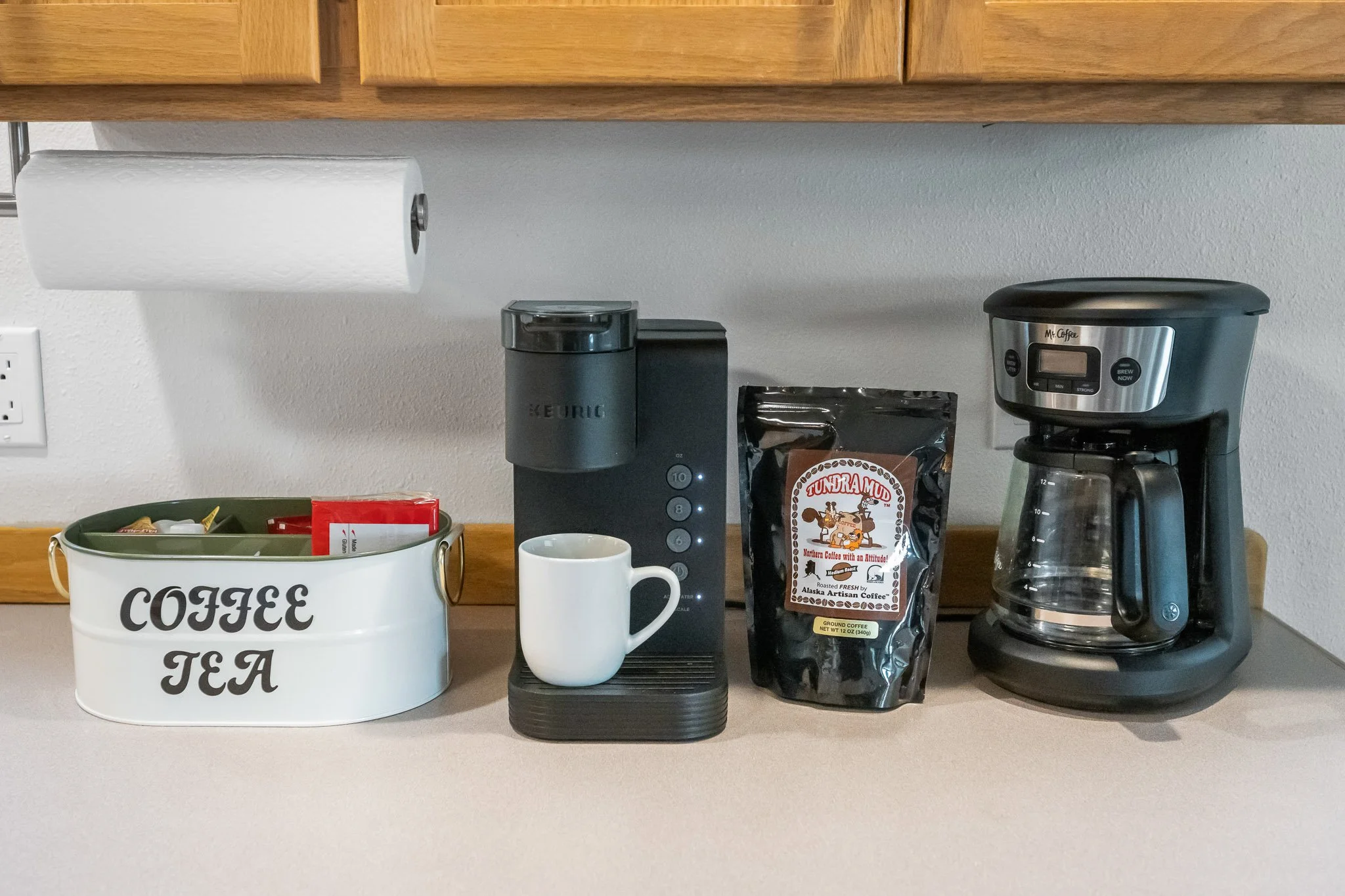 Kitchen countertop with coffee-making items: a white container with 'COFFEE TEA' printed on it, a white mug, a black coffee grinder, a bag of Tundra Mud coffee, a drip coffee maker, and a wooden cabinet above.