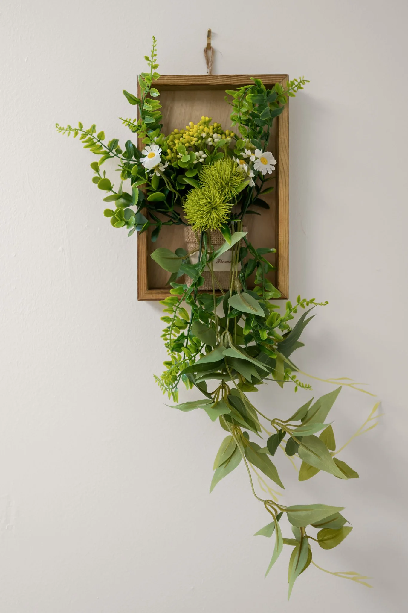 Wall-mounted wooden frame containing artificial green foliage, white daisies, and small green buds, with some greenery cascading downward.