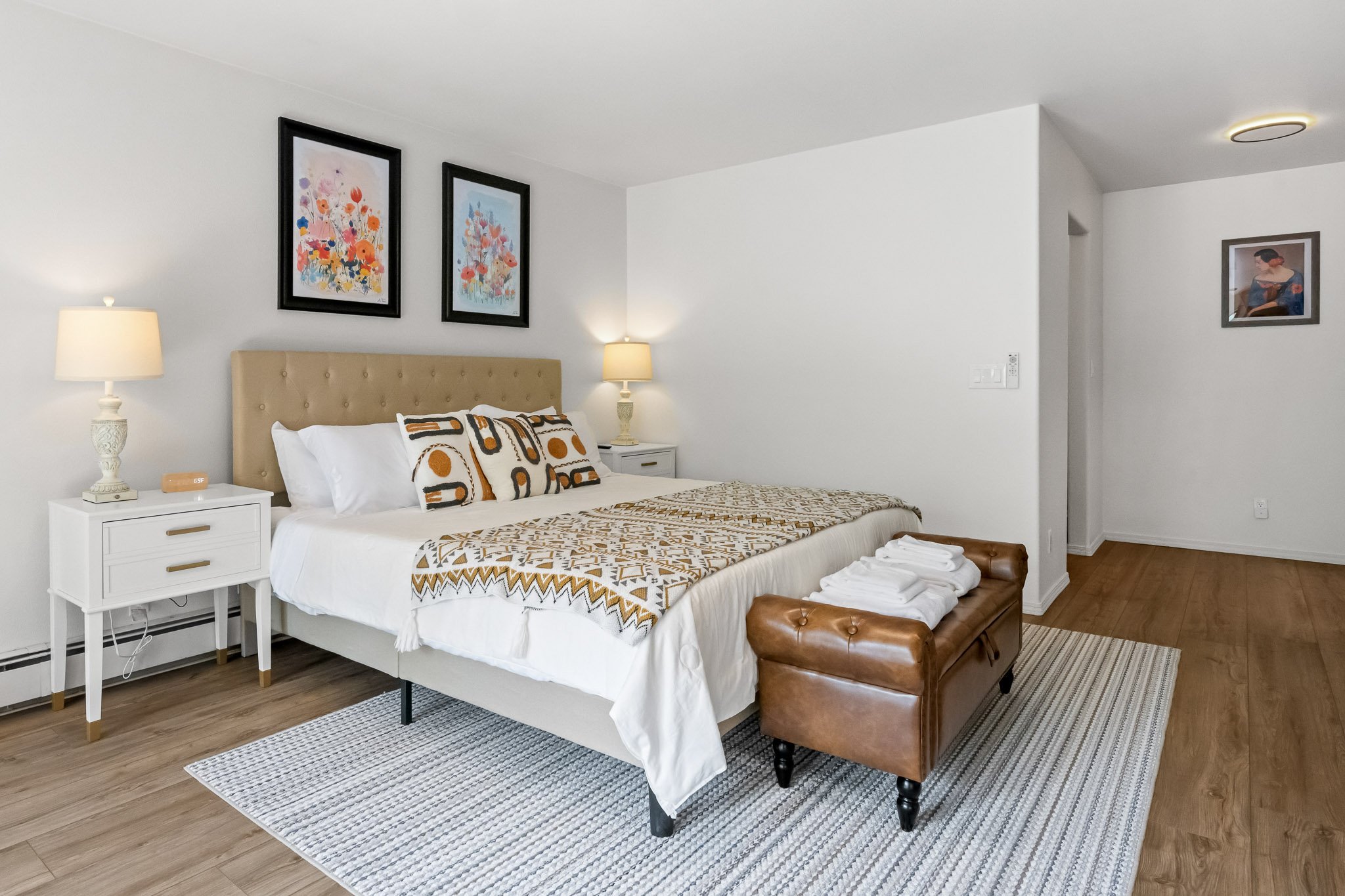 A bedroom with a bed, nightstands, lamps, artwork, and a leather bench at the foot of the bed, with wood flooring and a striped rug.