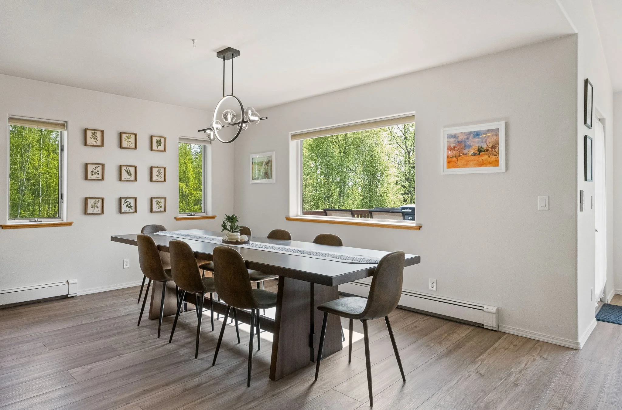 A modern dining room with a rectangular table surrounded by eight chairs, large windows showing green trees outside, and framed artwork on white walls, with a contemporary chandelier hanging from the ceiling.