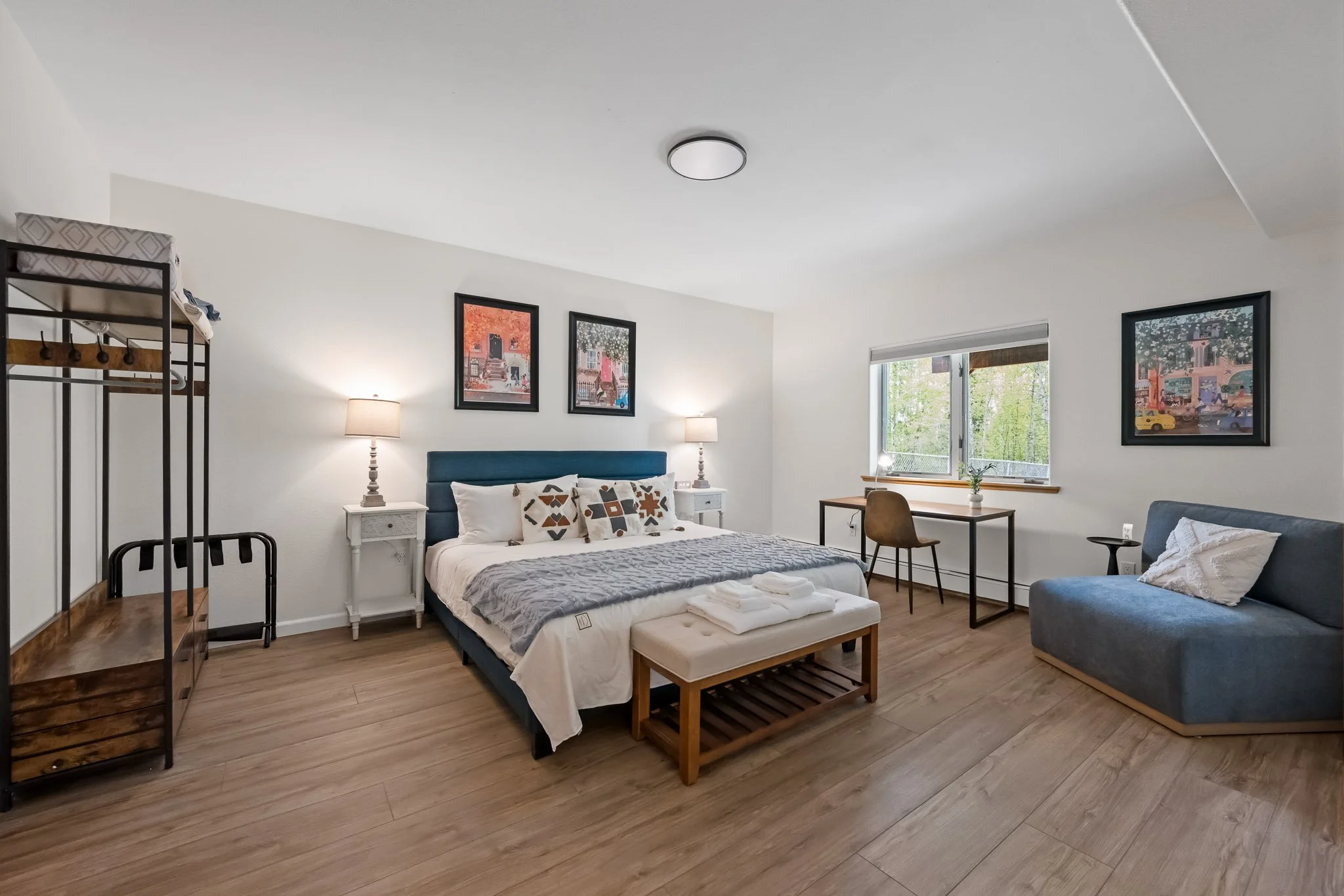 A bright, modern bedroom with a large bed, nightstands with lamps, framed artwork on the wall, a window with greenery outside, a desk and chair, a gray sofa with a pillow, and light wood flooring.
