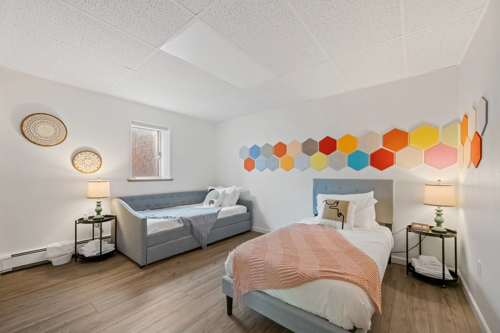 A bedroom with two beds, one with a peach quilt and another with a grey headboard and white bedding. Two nightstands with lamps, a window with frosted glass, and a wall decorated with colorful hexagon tiles.