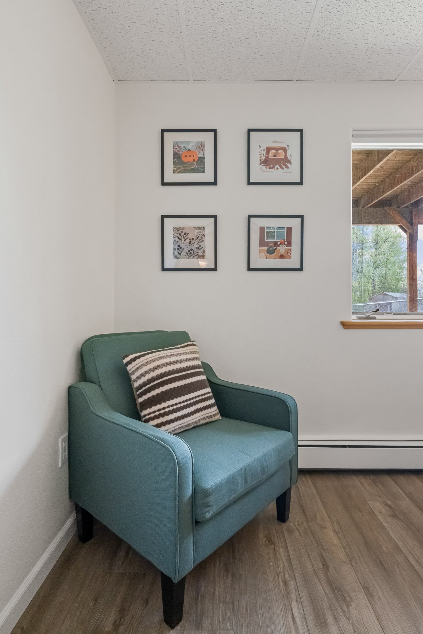 A teal upholstered armchair with dark wooden legs and a black and white striped throw pillow, positioned against white walls and a wooden floor, with a window showing a view of green trees and a wooden outdoor structure, and four framed pictures on t